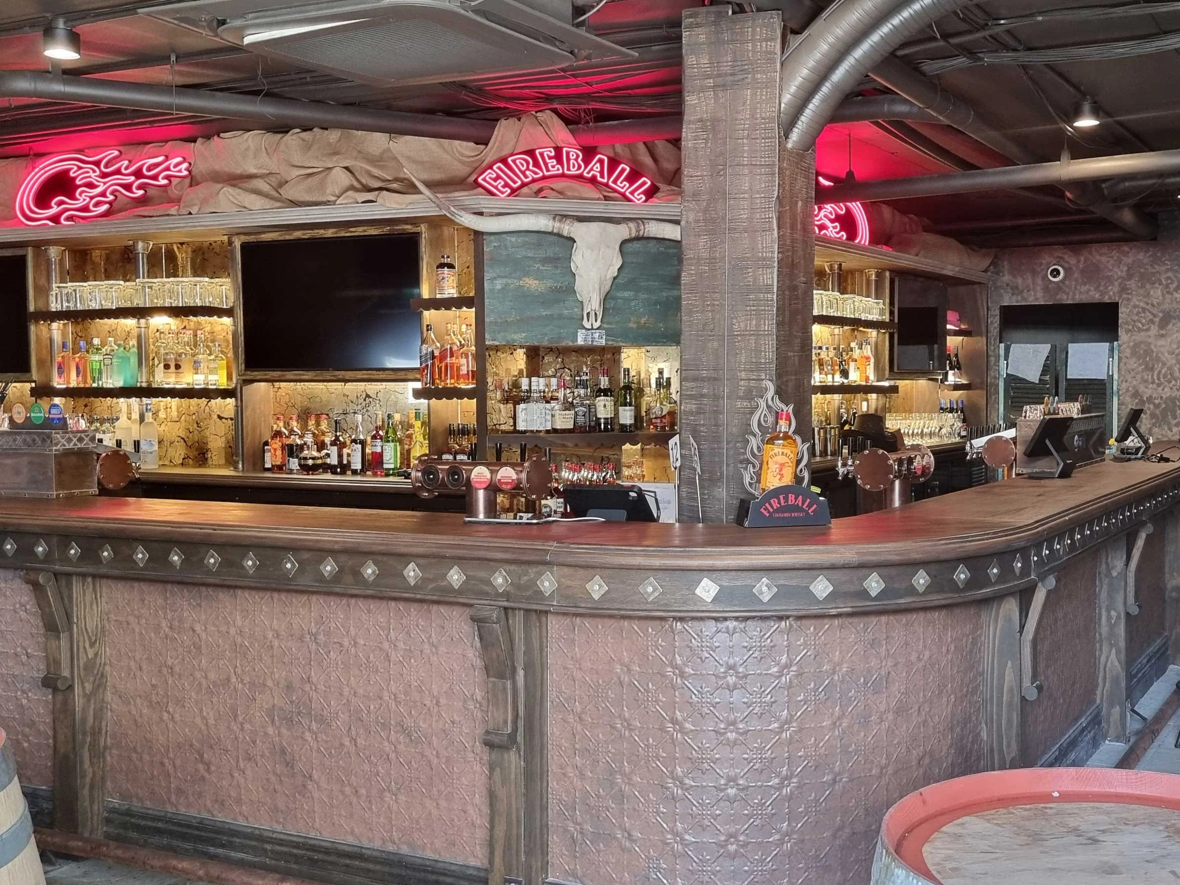Empty bar counter with liquor bottles on shelves, fireplace sign, neon Fireball sign, skull mounted above bar, cash registers, pointing hand sign, and decorative elements in a rustic style.