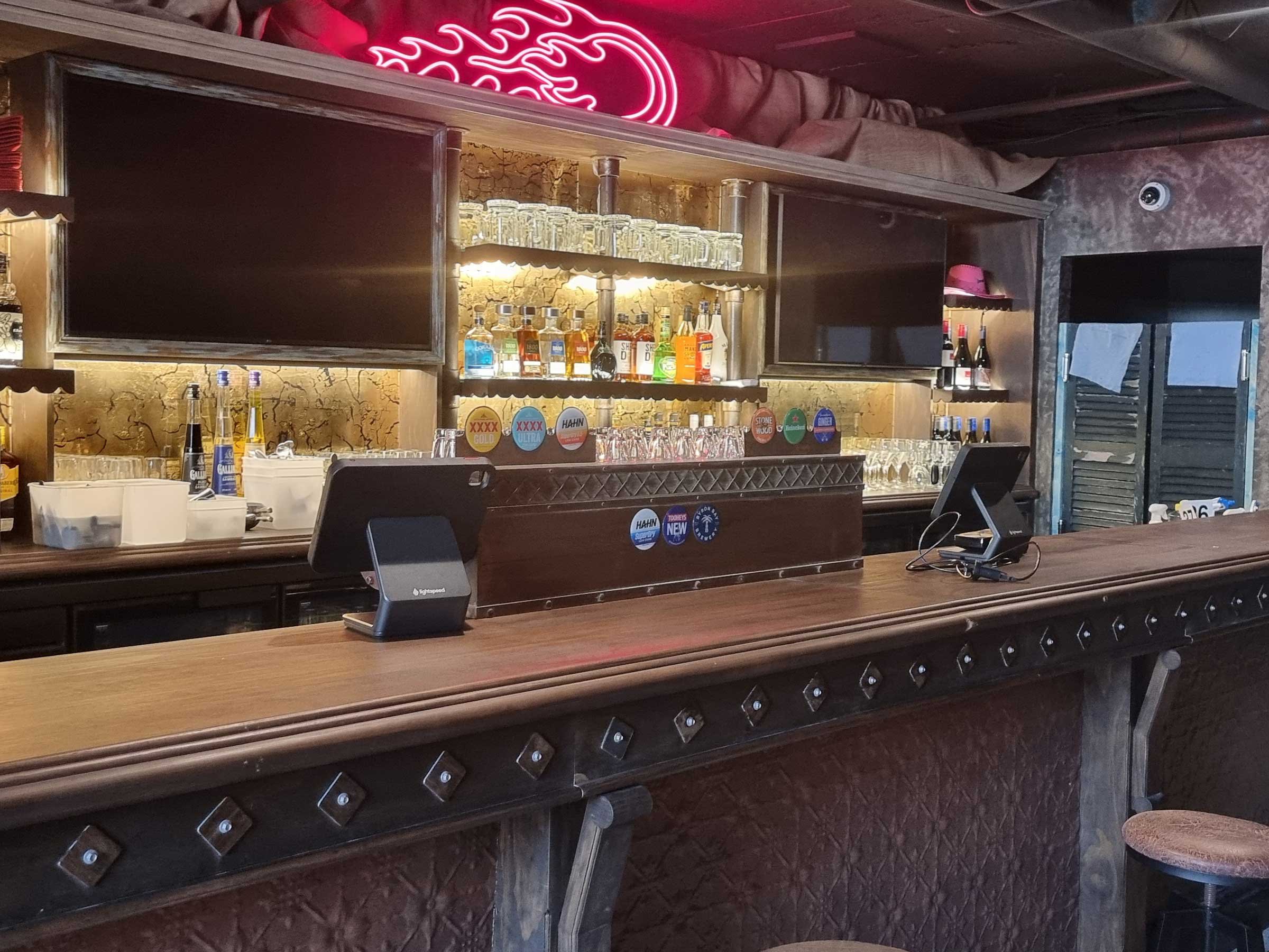 Empty bar with liquor bottles and glasses, cash registers, and neon signs inside a dimly lit bar or pub.