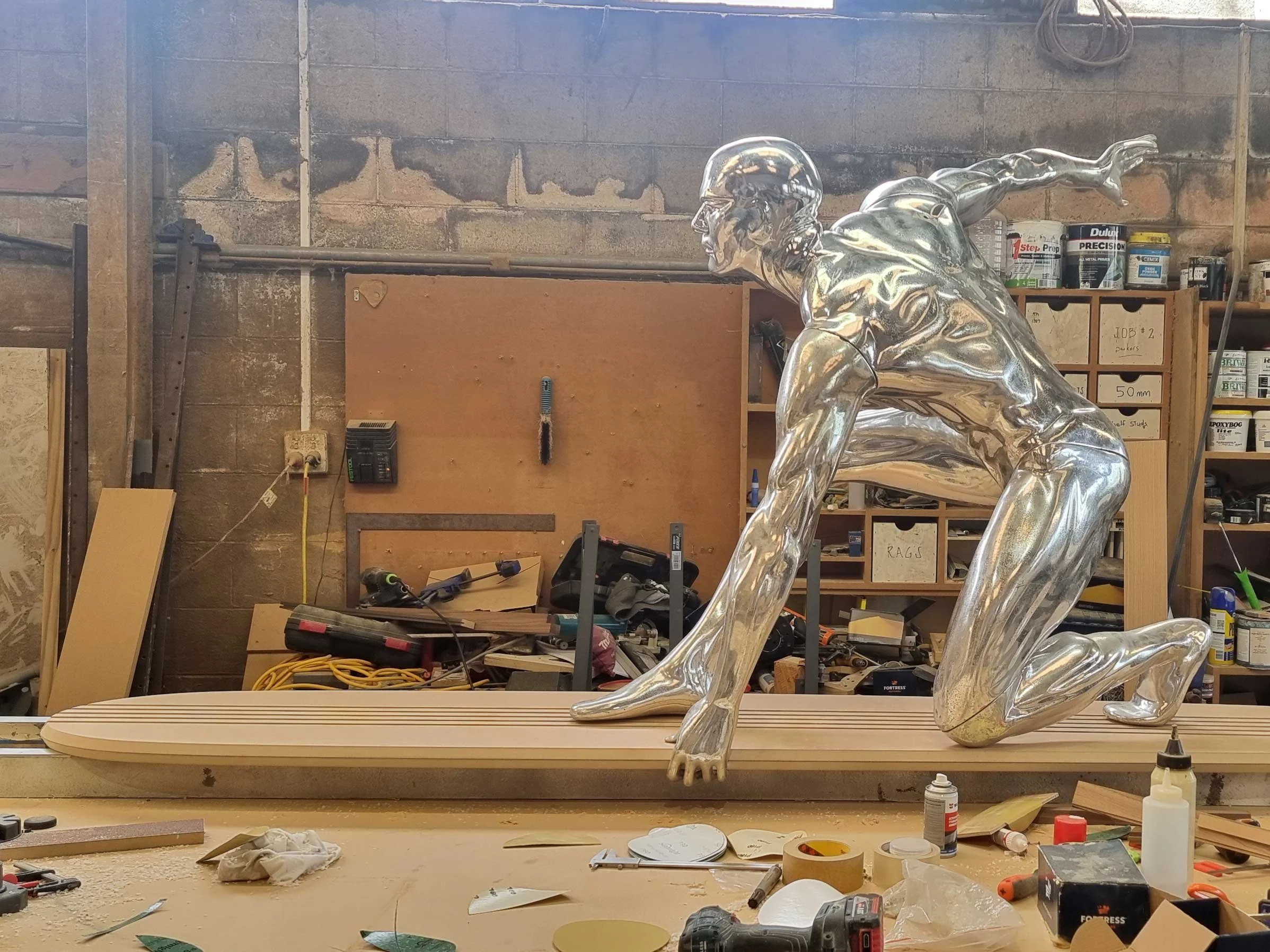 A reflective metallic sculpture of a person on a skateboard in a woodworking workshop.