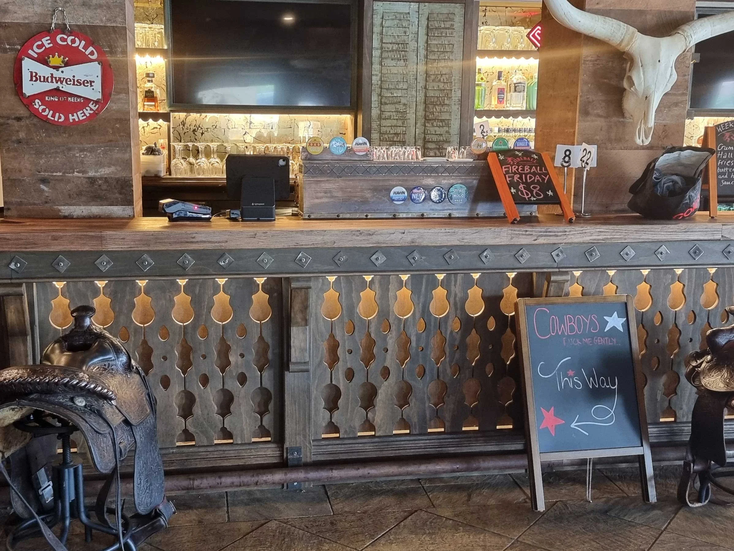Bar counter with a chalkboard sign pointing to Cowboys ride and a nearby horse saddle. Behind the counter, bottles of alcohol on shelves and bar equipment are visible. A cow skull hangs on the wall, and a round Budweiser sign is mounted on the wall.