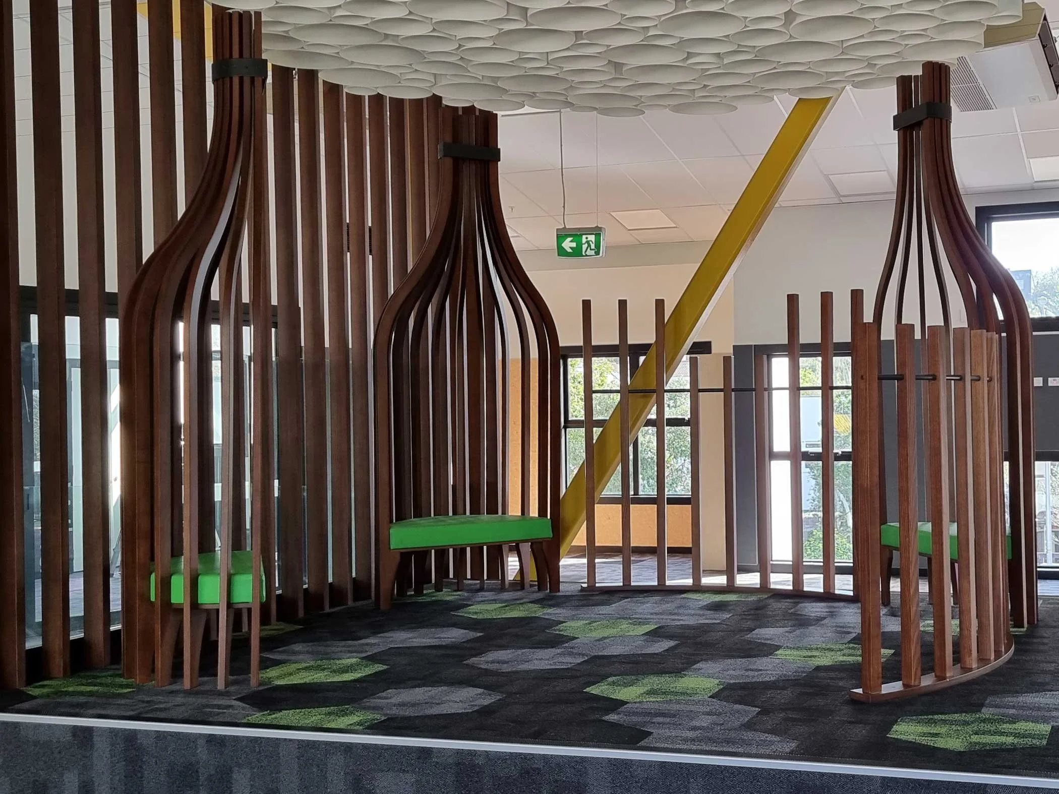 Indoor playground with curved wooden slat structures, green cushioned seats, and a yellow slide, on a black and green hexagon-patterned carpet.
