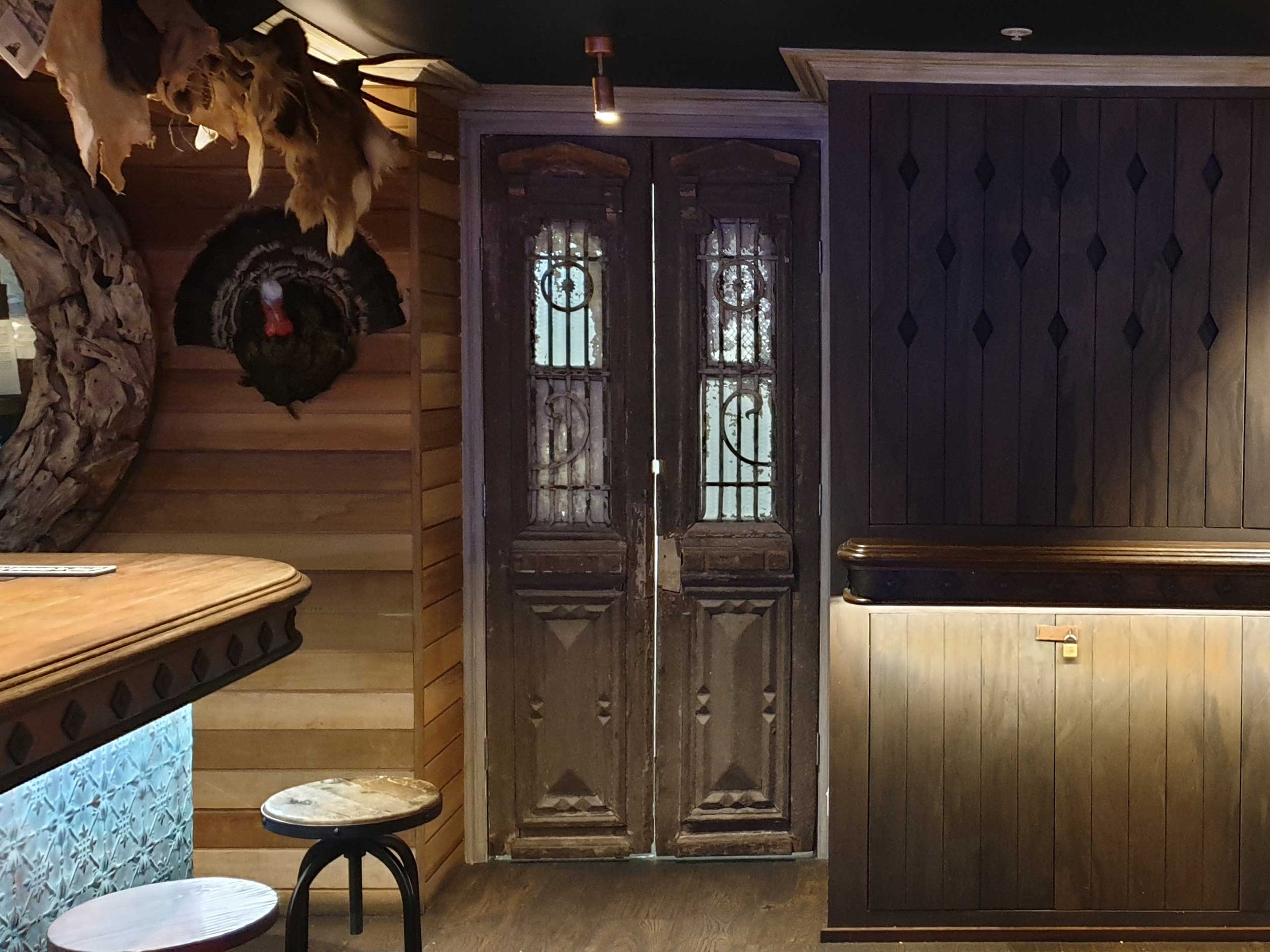 Interior of a rustic bar or restaurant with wooden walls, a bar counter with a decorative front, a mounted turkey head on the wall, and a set of closed wooden doors with glass and wrought iron details.