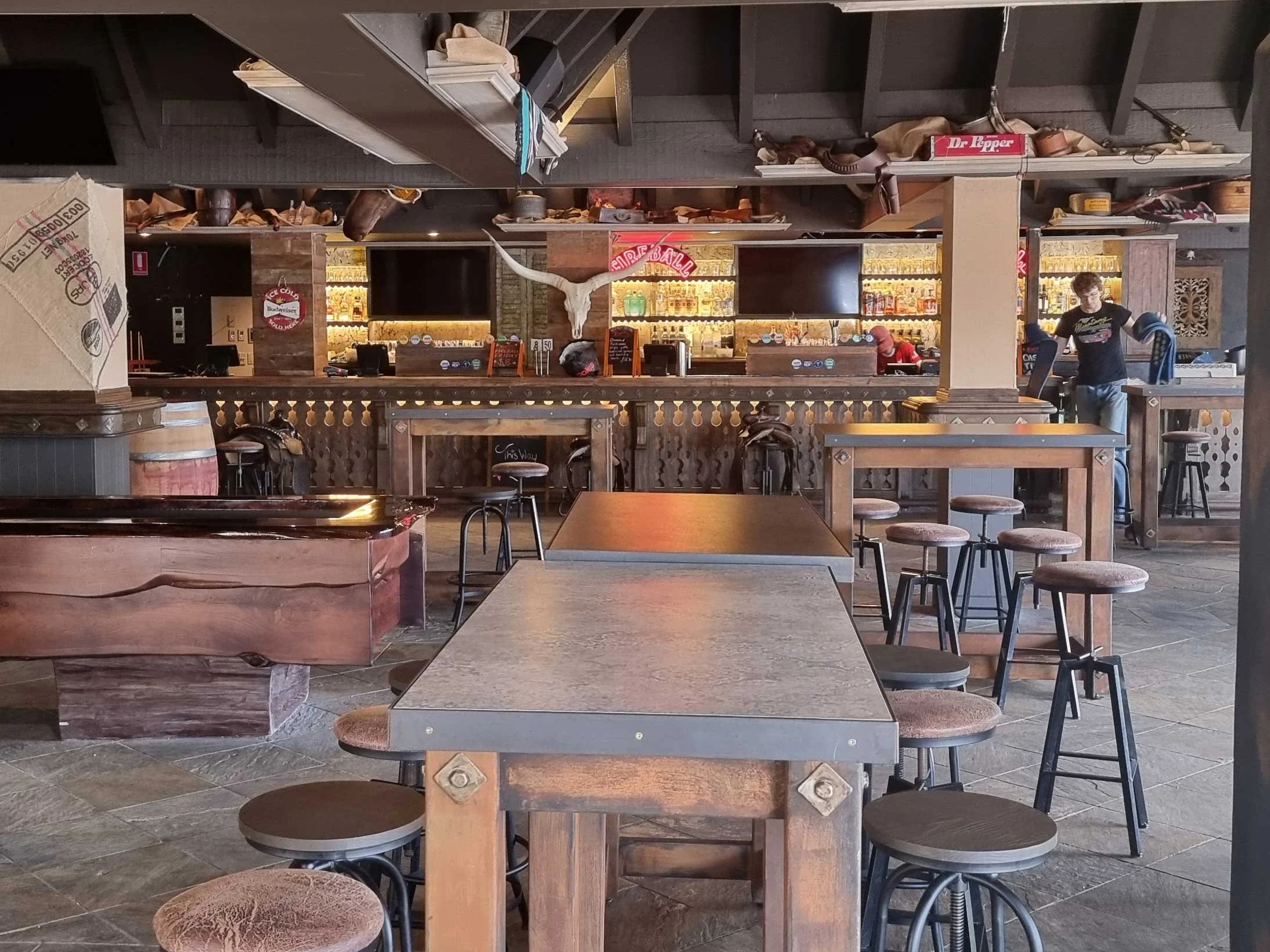Interior of a rustic bar with wooden tables and bar stools, a bar counter in the background with bottles and a mounted cow skull on the wall.