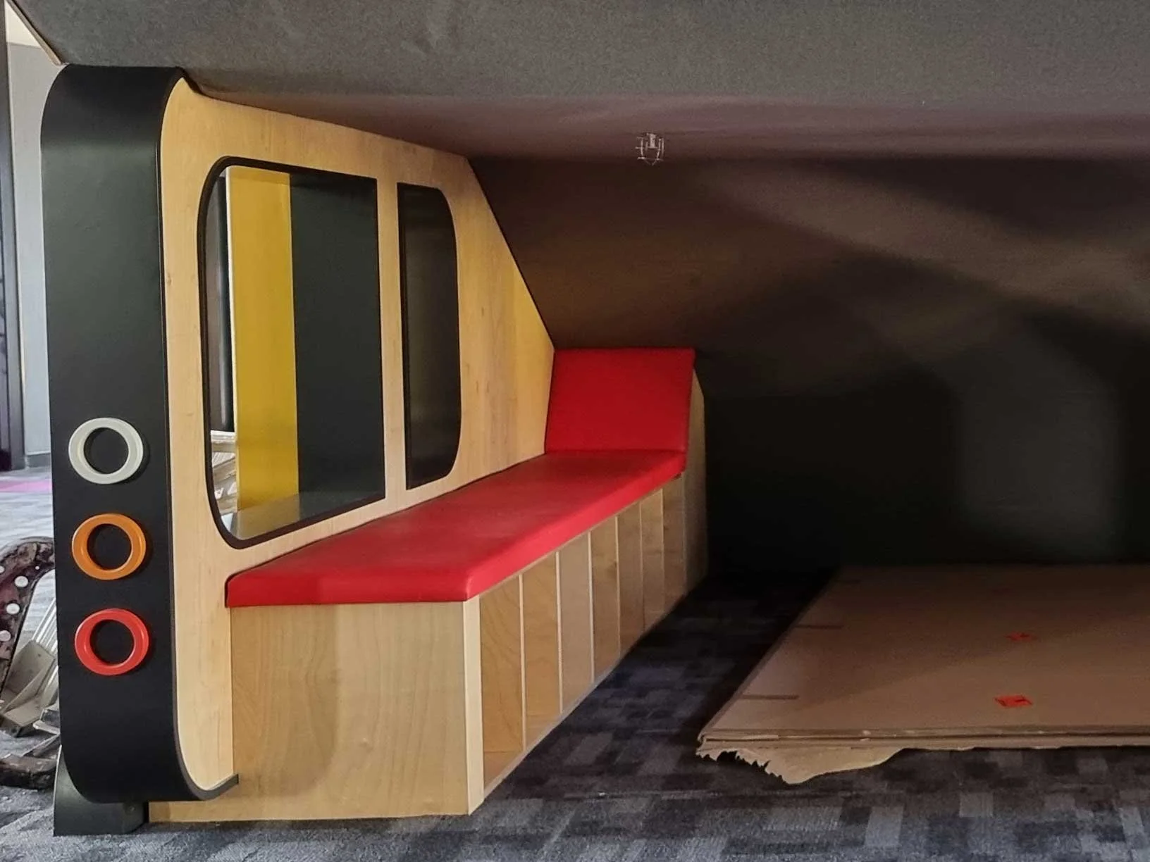 A modern wooden bench with a red padded seat, a black armrest with four colorful circular holes, and a mirror or glass panel on top, positioned against a dark wall with a stack of cardboard underneath.
