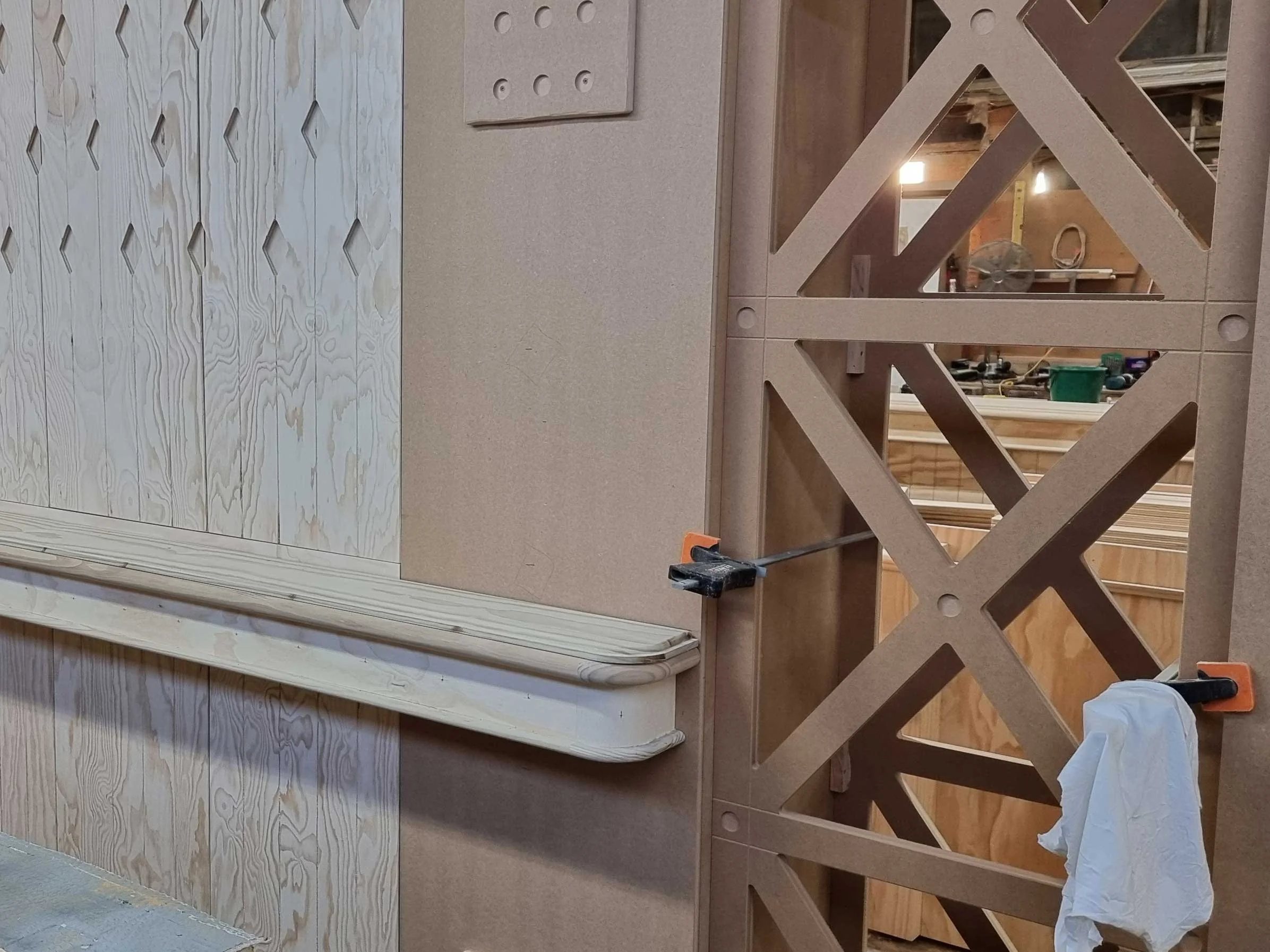 Interior view of a construction site with unfinished drywall, a wooden trim, and a decorative wooden partition with geometric cut-outs. Tools and construction materials are visible in the background.