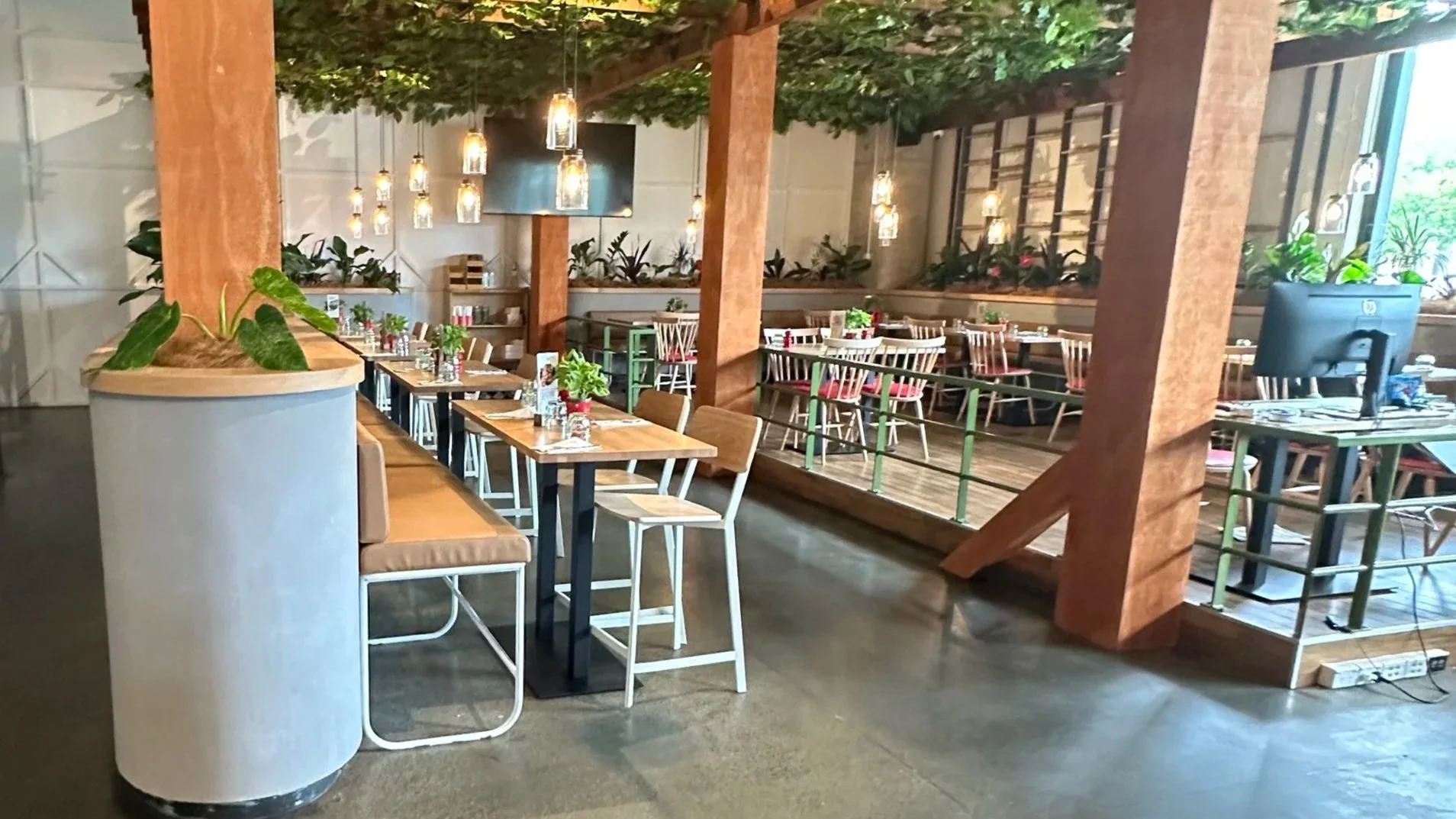 Interior of a modern restaurant with a mix of wooden and metal furniture, potted plants, hanging pendant lights, and a raised dining area with a green railing.