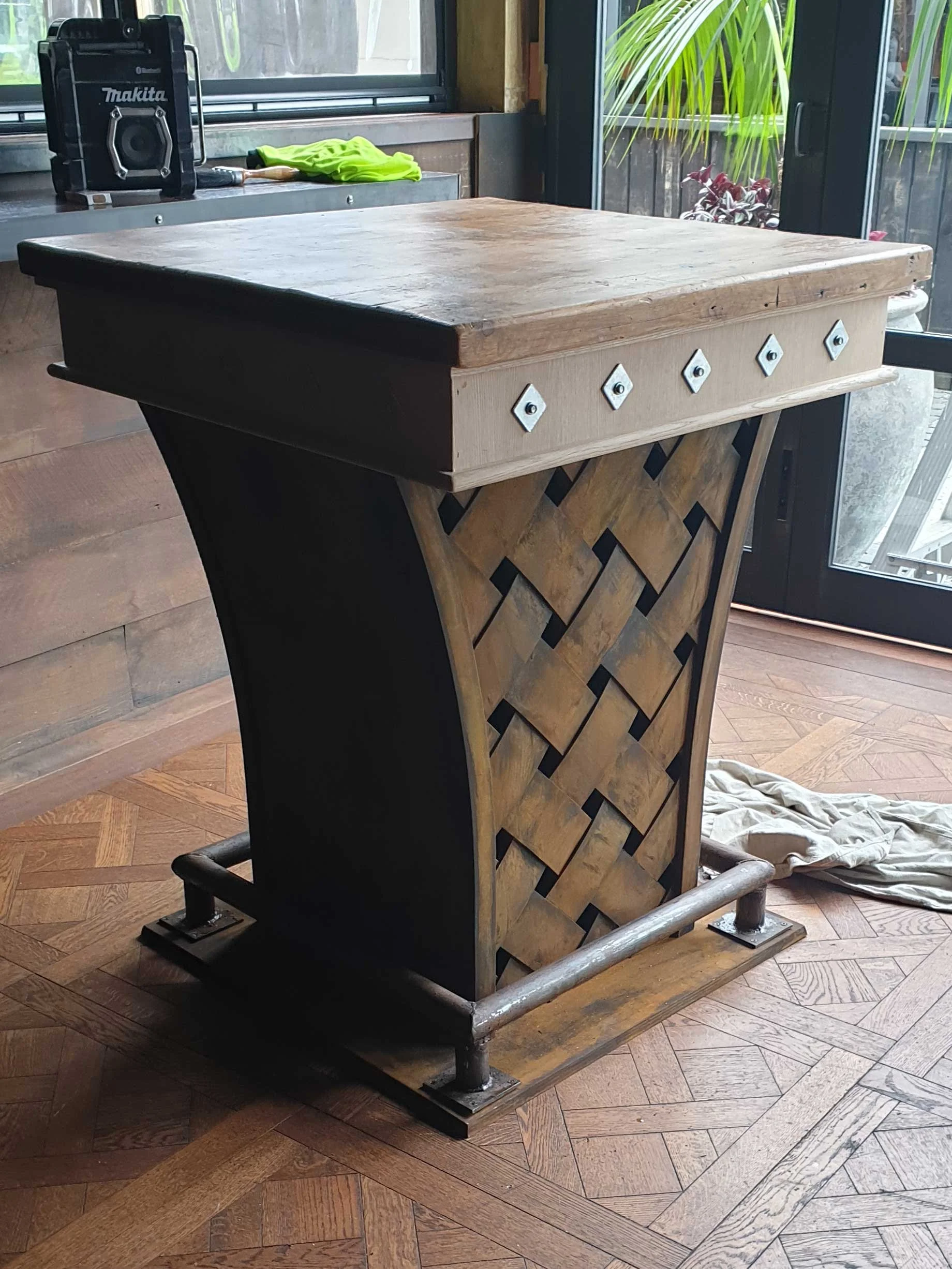 A wooden bar counter with a patterned front, metal studs, and a wooden top, situated on a parquet wood floor near large windows with plants outside.