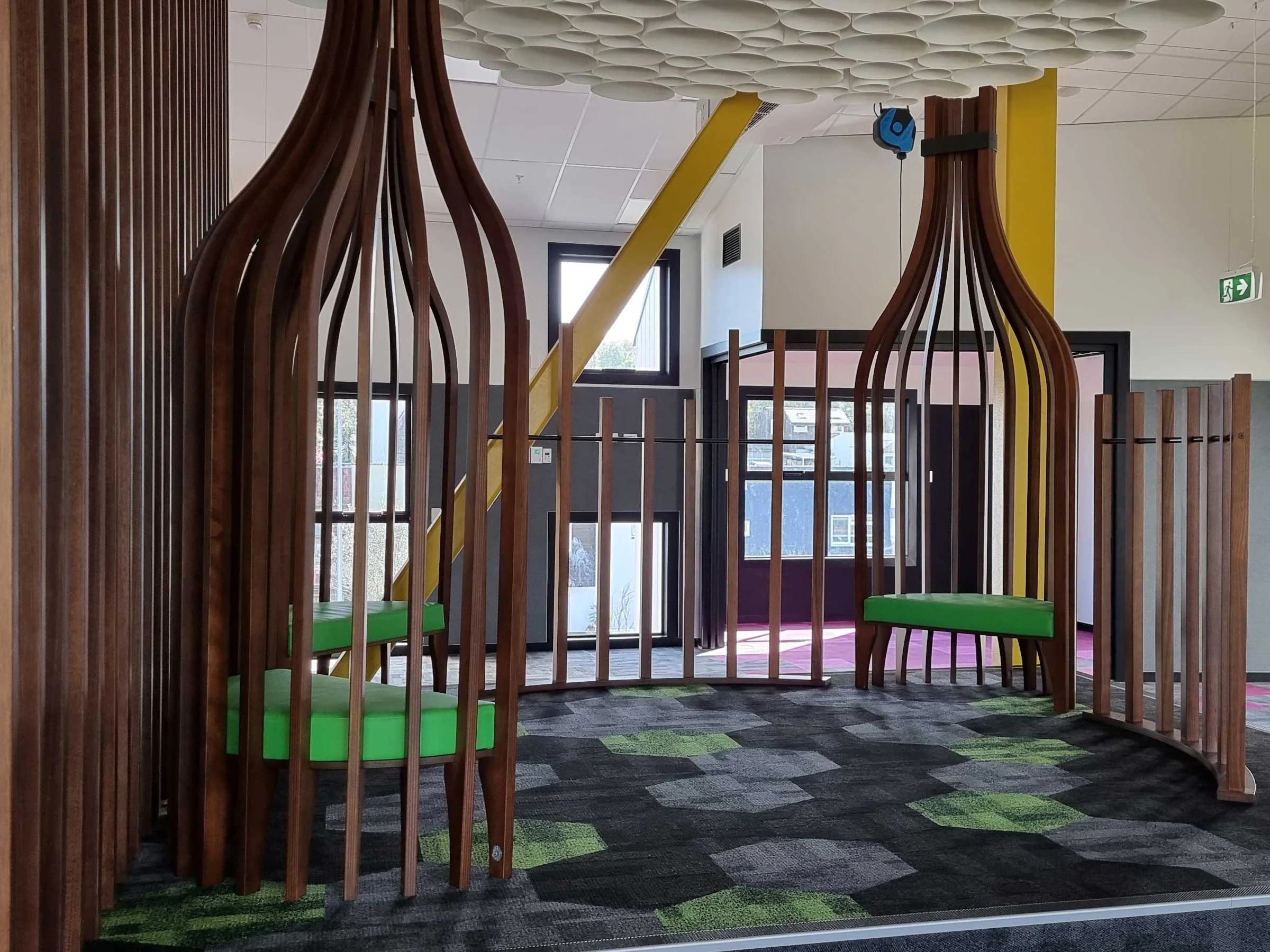 Modern indoor play area with wooden structures, green cushioned seats, and a patterned carpet. Large windows provide natural light.