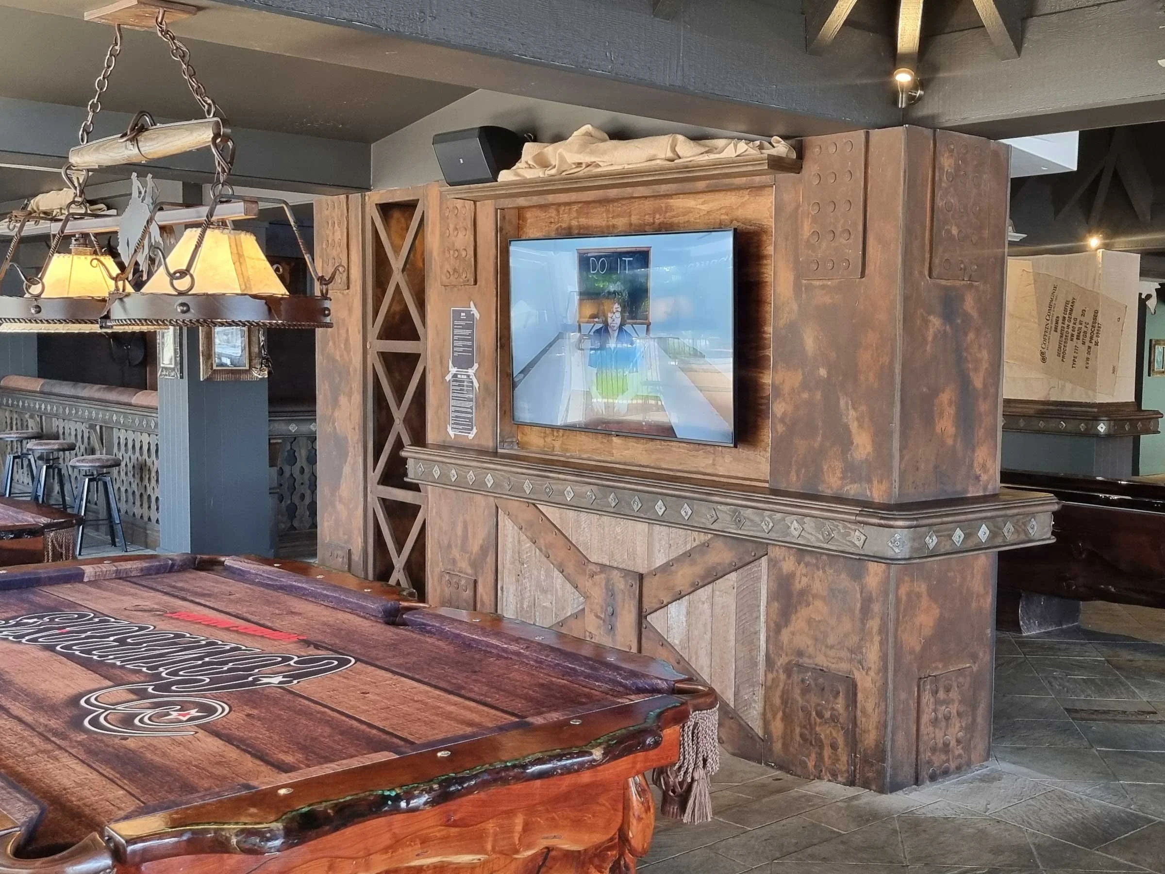 Interior of a rustic-themed bar with a wooden pool table, a mounted television on a wooden partition, and a chandelier overhead.