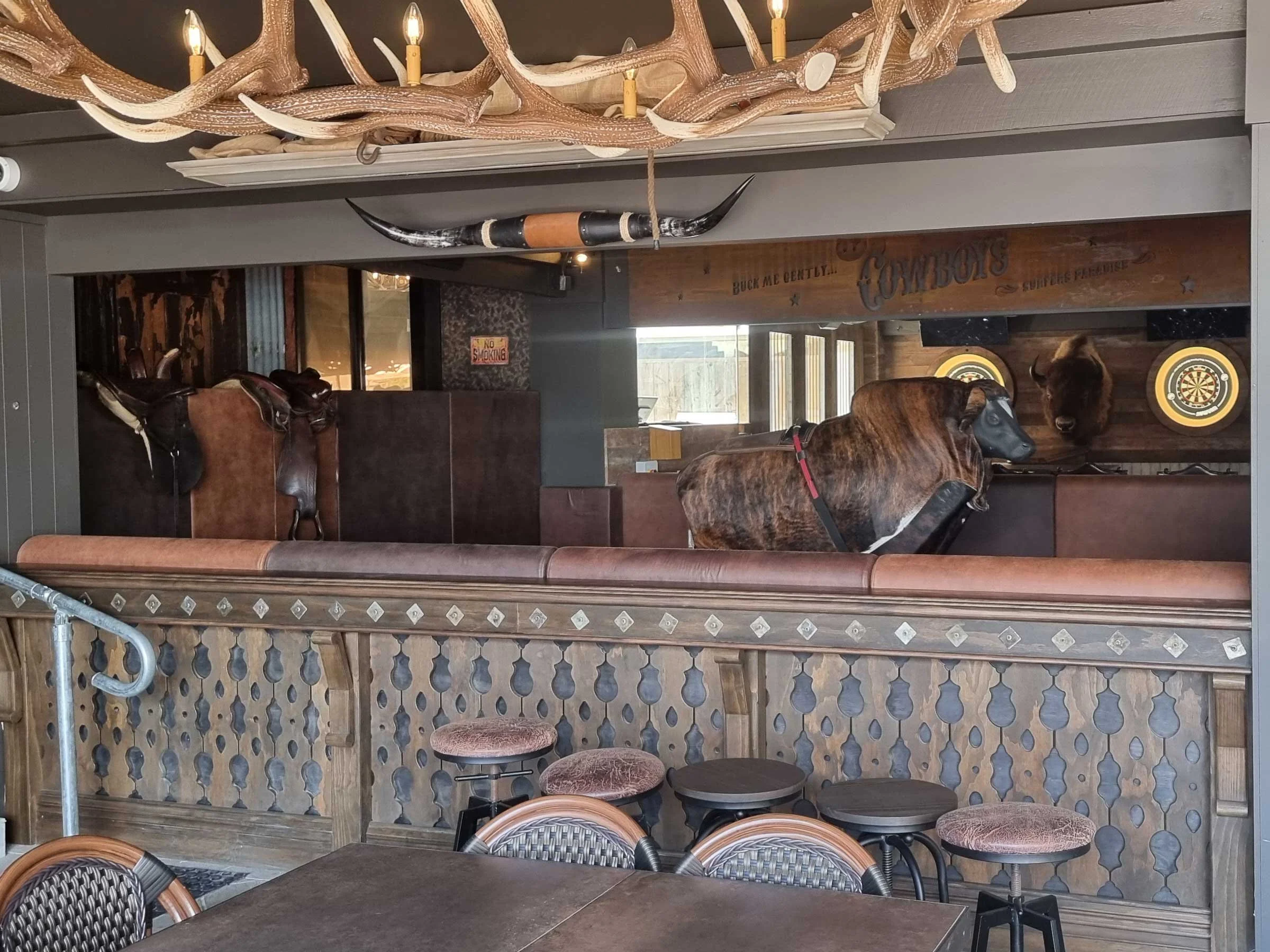 Interior of a western-themed bar featuring a wooden railing, bar stools with upholstered seats, a hanging antler chandelier with candle-shaped lights, a brown and black bison head mounted on the wall, a saddle on the railing, and a dartboard in the b