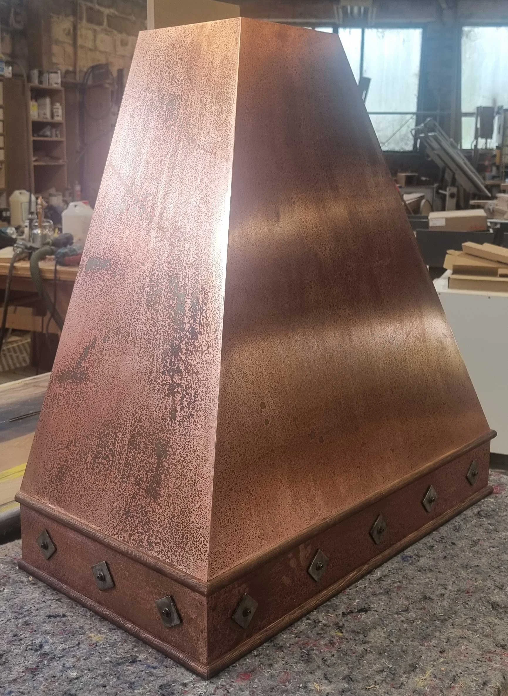 A large, metallic, copper-colored pyramid-shaped object with a textured surface, resting on a workbench in a woodworking shop.