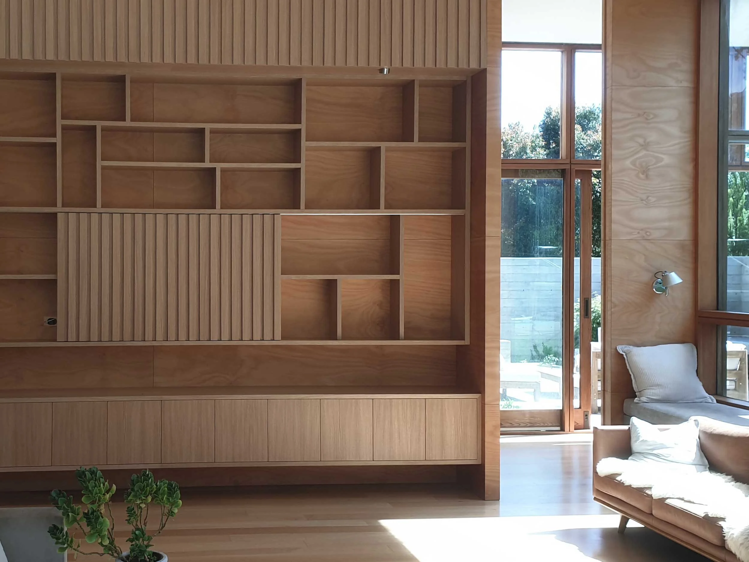Interior of a modern living room with a wooden built-in bookcase, large windows, a beige sofa with pillows, and a small potted plant.