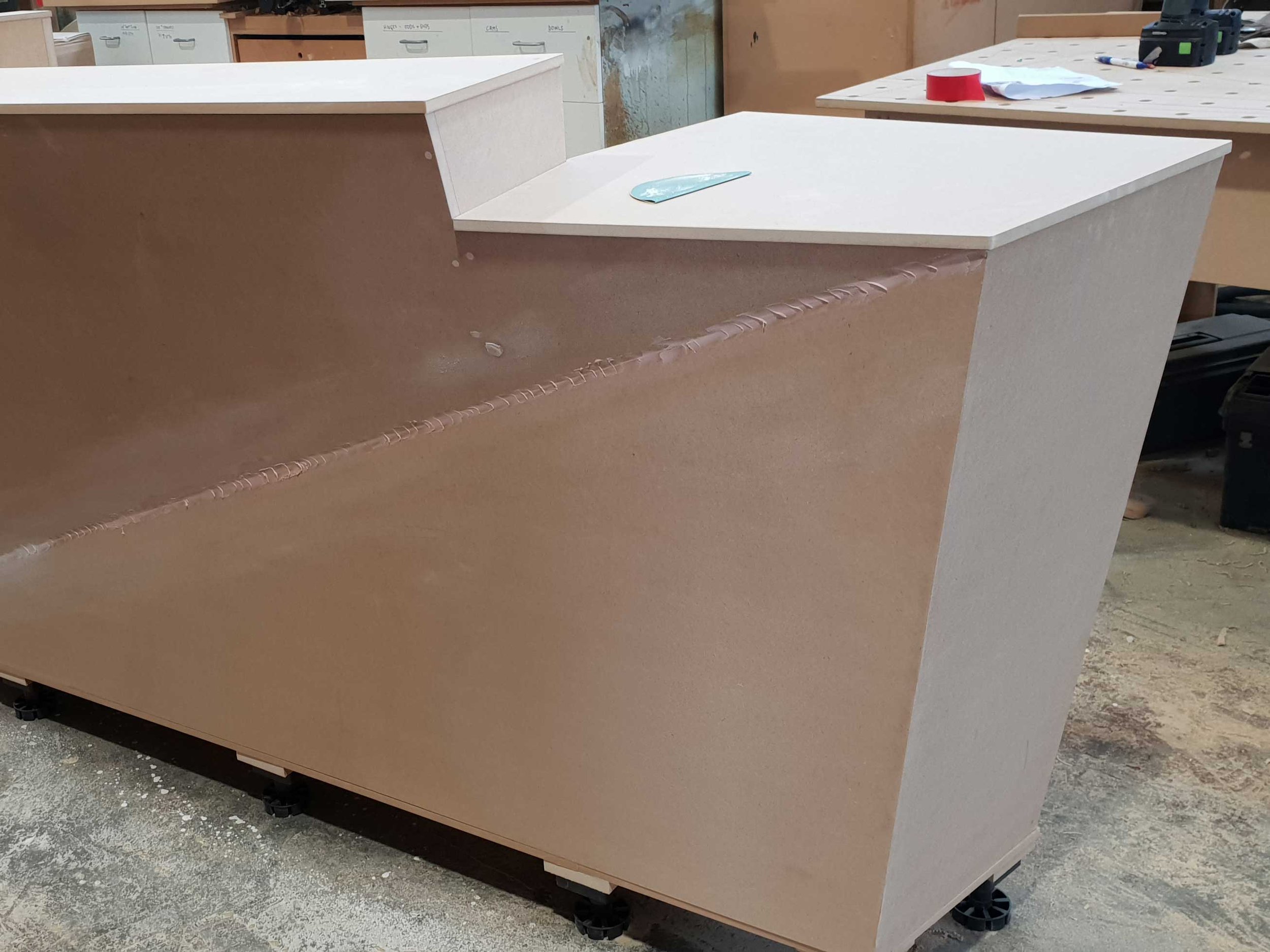 A partially assembled L-shaped kitchen or work counter with a white countertop and unfinished wooden panels, situated in a woodworking or construction workshop with tools, papers, and other materials visible in the background.
