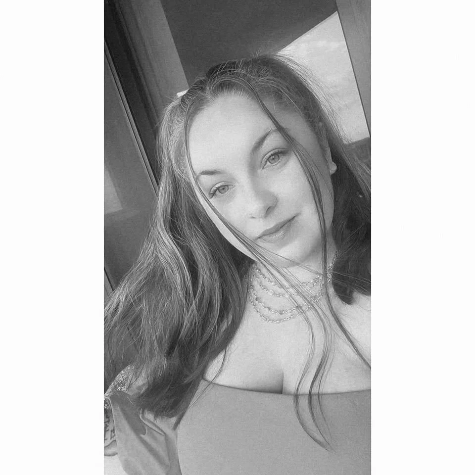 A woman with curly dark hair sits on a bench in a room with neutral-colored walls, with sunlight casting shadows.