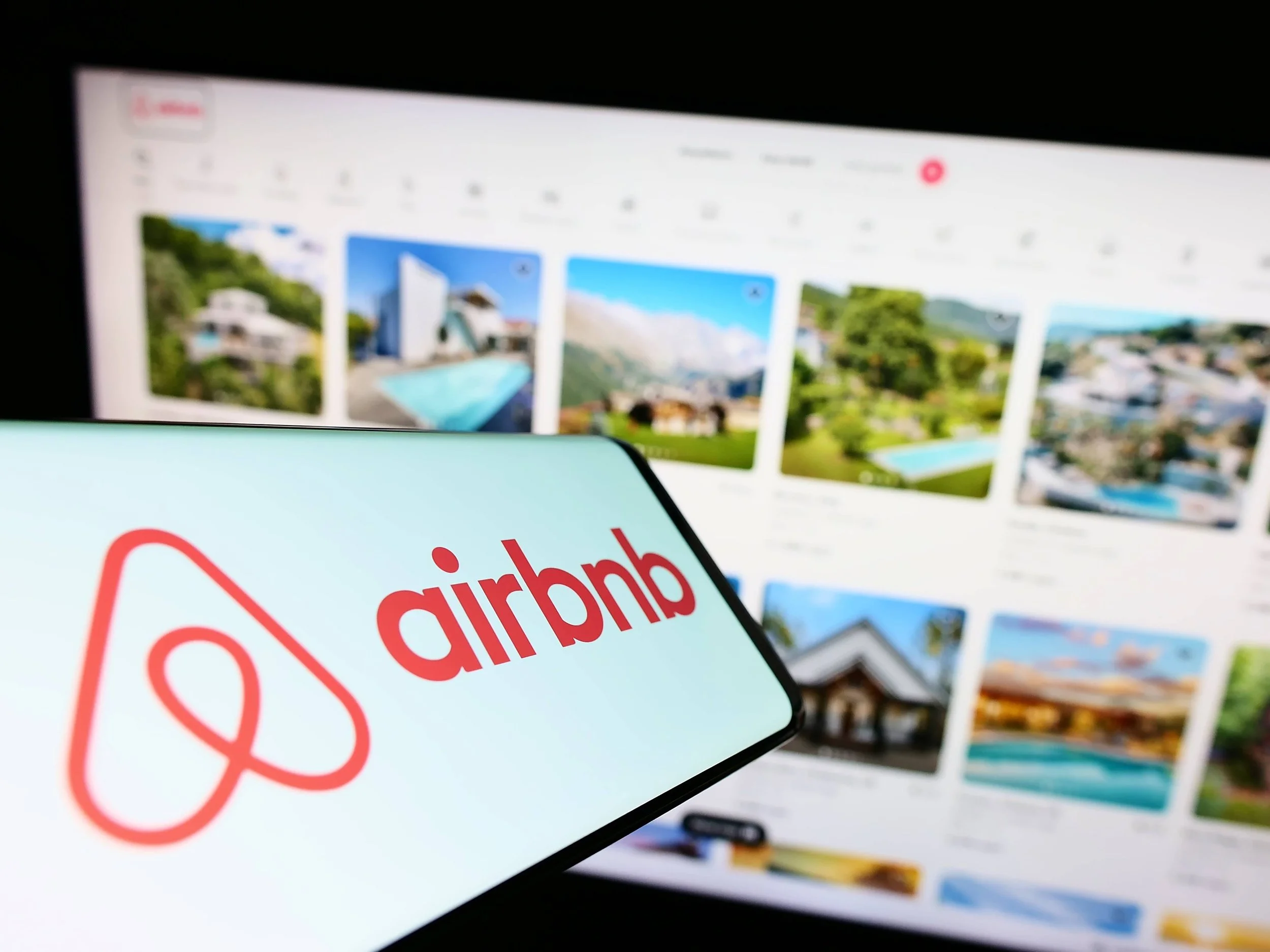 Smartphone displaying the Airbnb logo in front of a computer screen showing multiple vacation rental photos.
