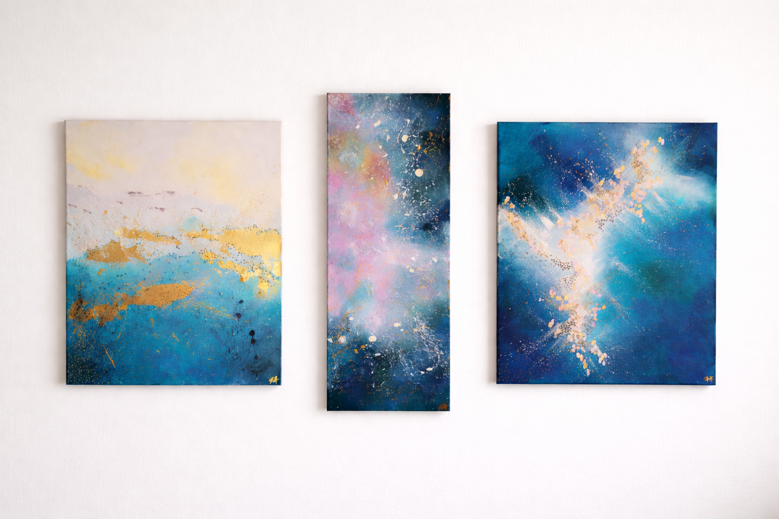 Three abstract paintings with splashes of blue, pink, gold, and white on a white gallery wall.