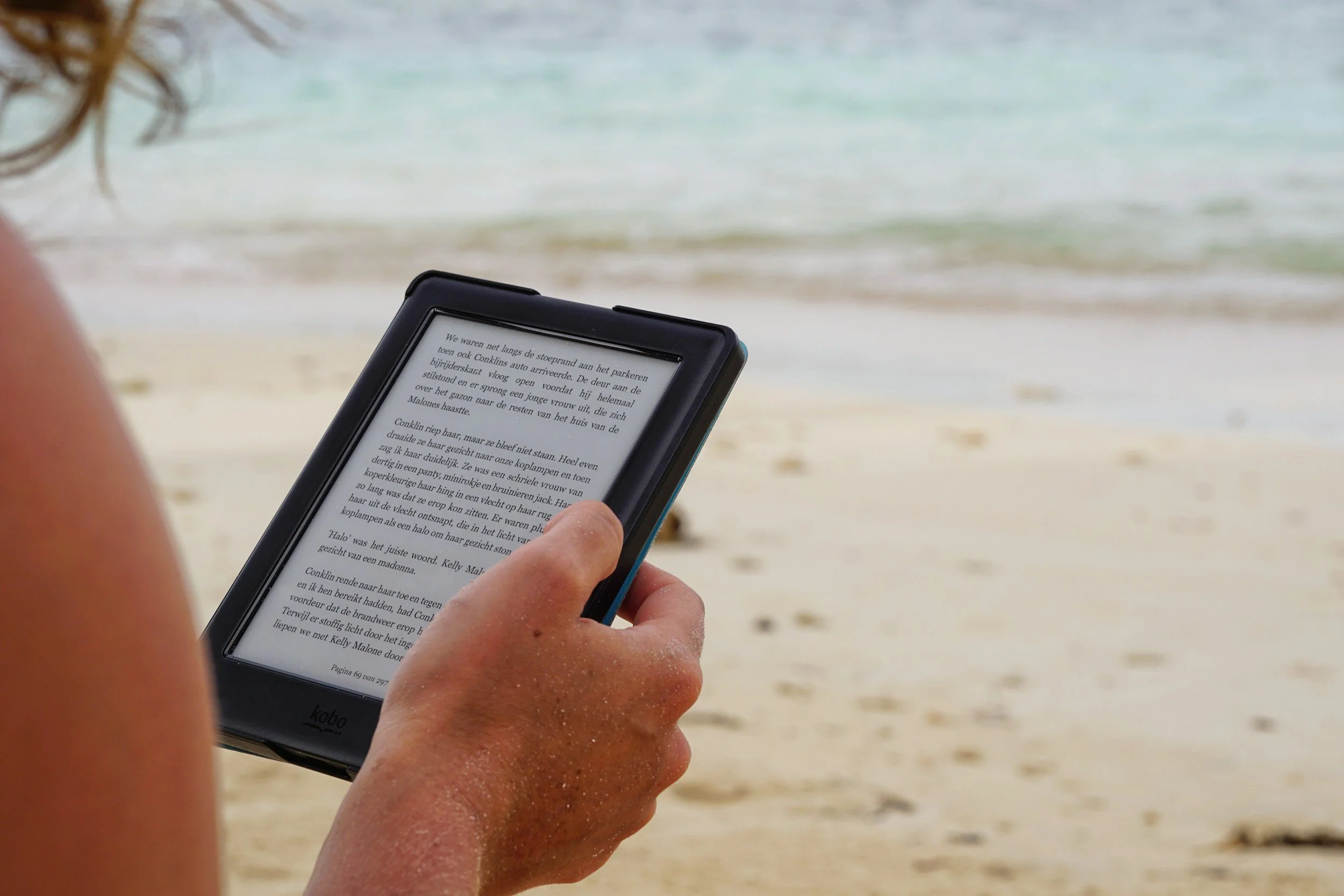 Person holding an e-reader on a sandy beach with the ocean in the background.