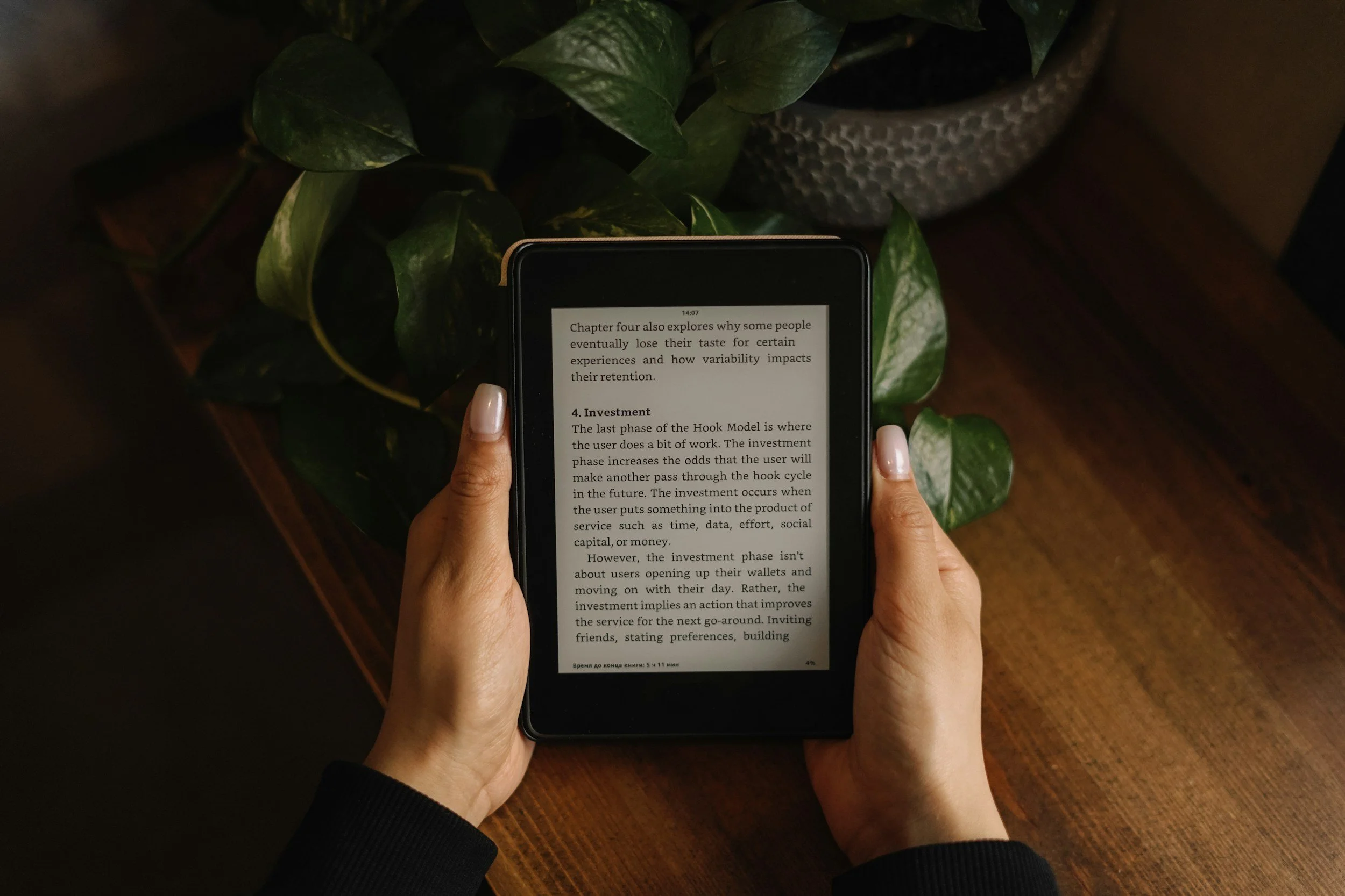 Person holding an e-reader displaying text about investment and the hook model, sitting at a wooden table near a potted plant.