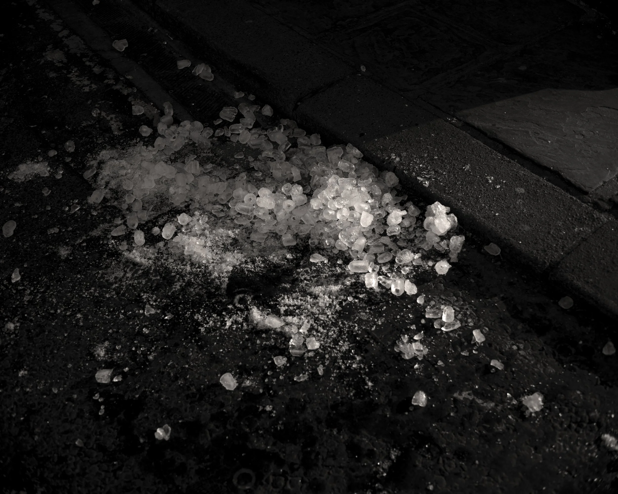 Salt crystals spilled on a dark pavement surface near a curb.