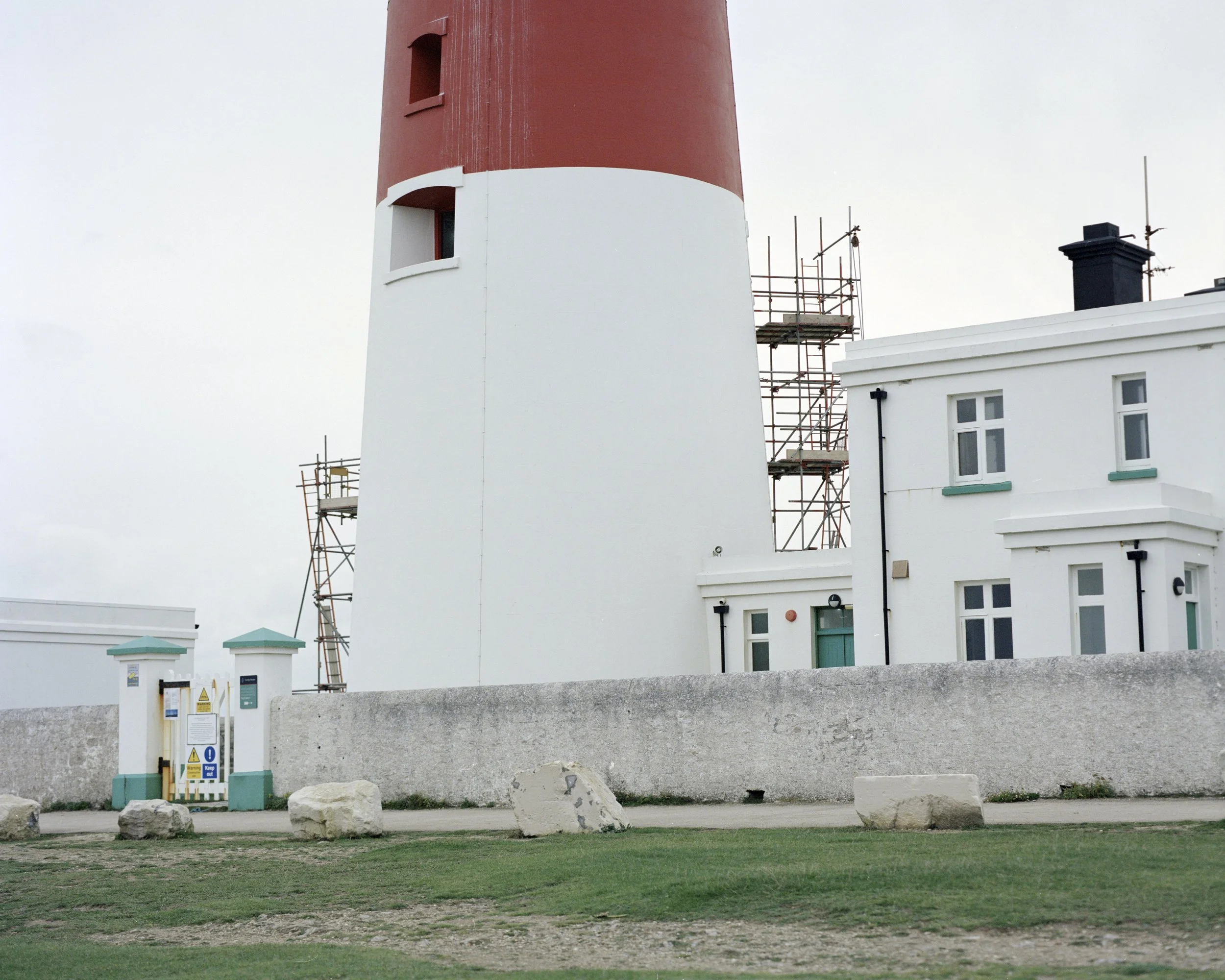 A lighthouse with a red top and white body near a white building, with scaffolding on the side, and a concrete wall in front surrounded by grass and rocks, under an overcast sky.
