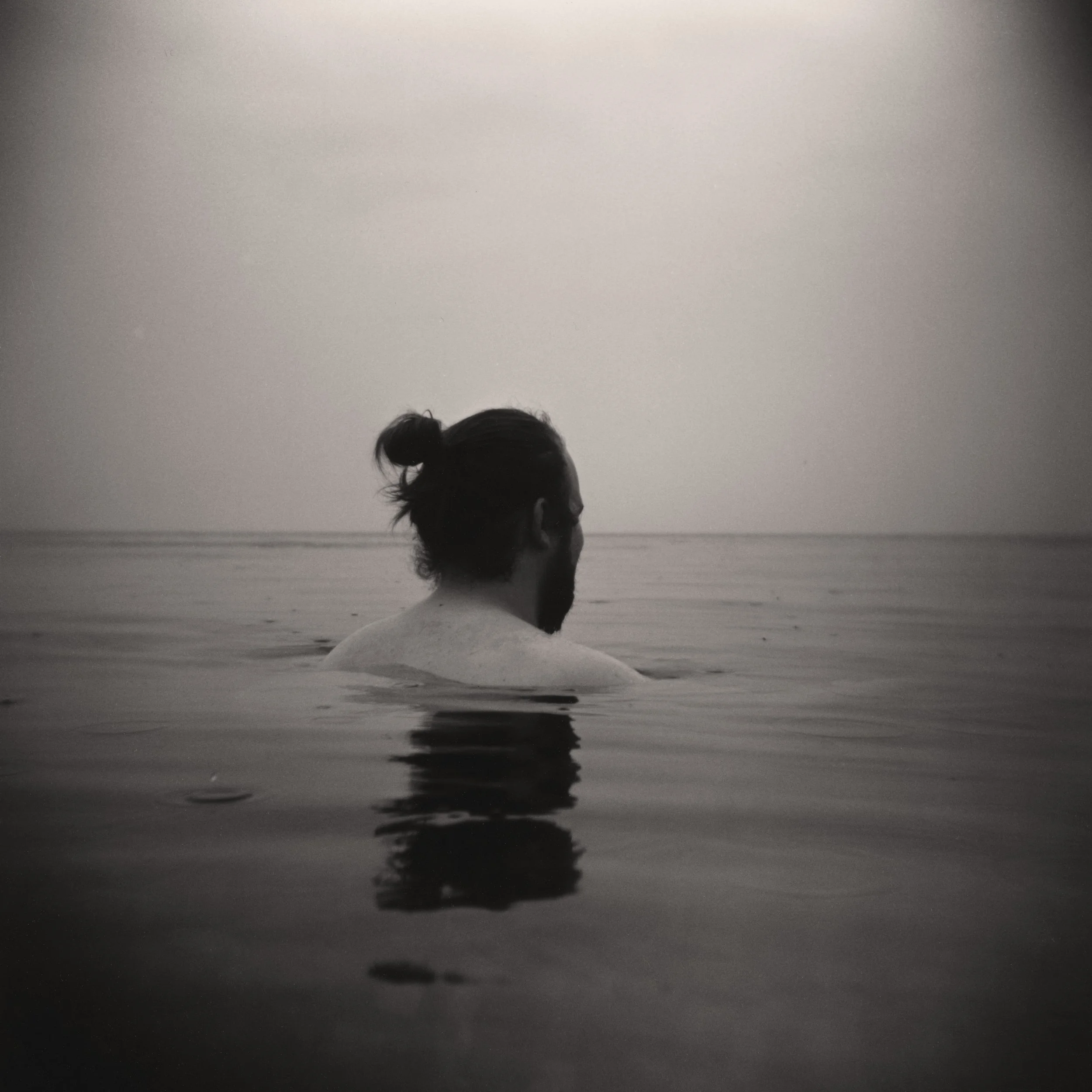 A black and white photo of a man with long hair in a bun, swimming in calm water, facing away from the camera with the horizon in the background.