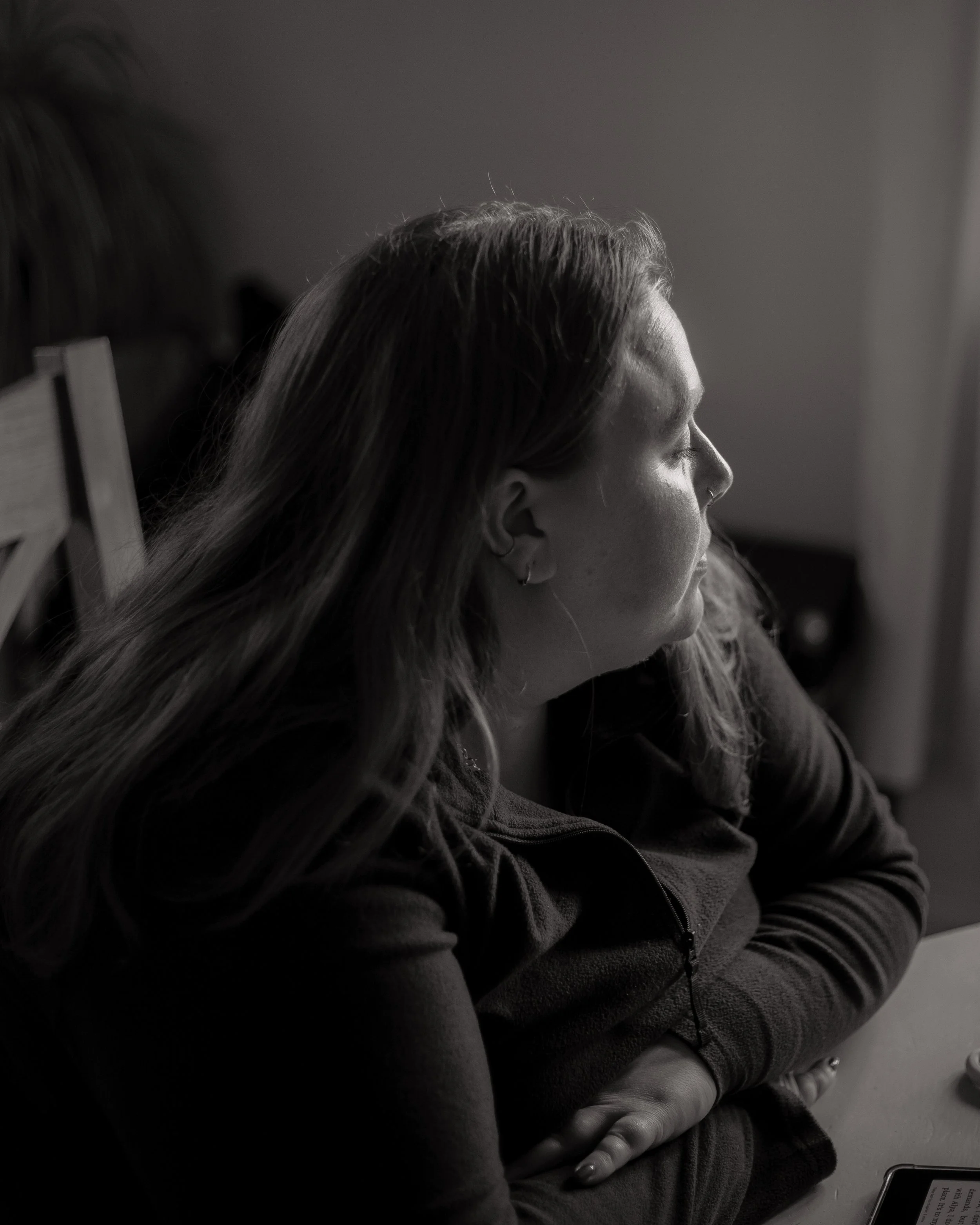 A woman with long hair and earrings seated with arms crossed, wearing a zip-up jacket, facing to the right in a black and white photo.