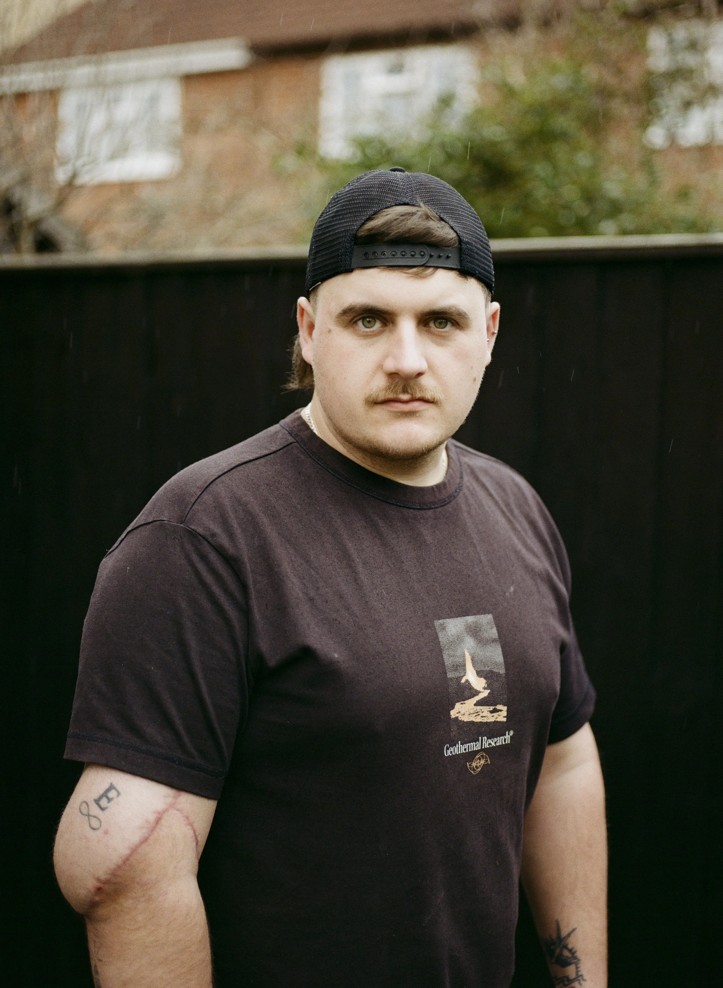 A young man with blonde hair, green eyes, and tattoos on his arms, wearing a black baseball cap backwards and a black T-shirt with a graphic design and the text 'Geothermal Research,' standing outdoors in front of a blurred background that includes a