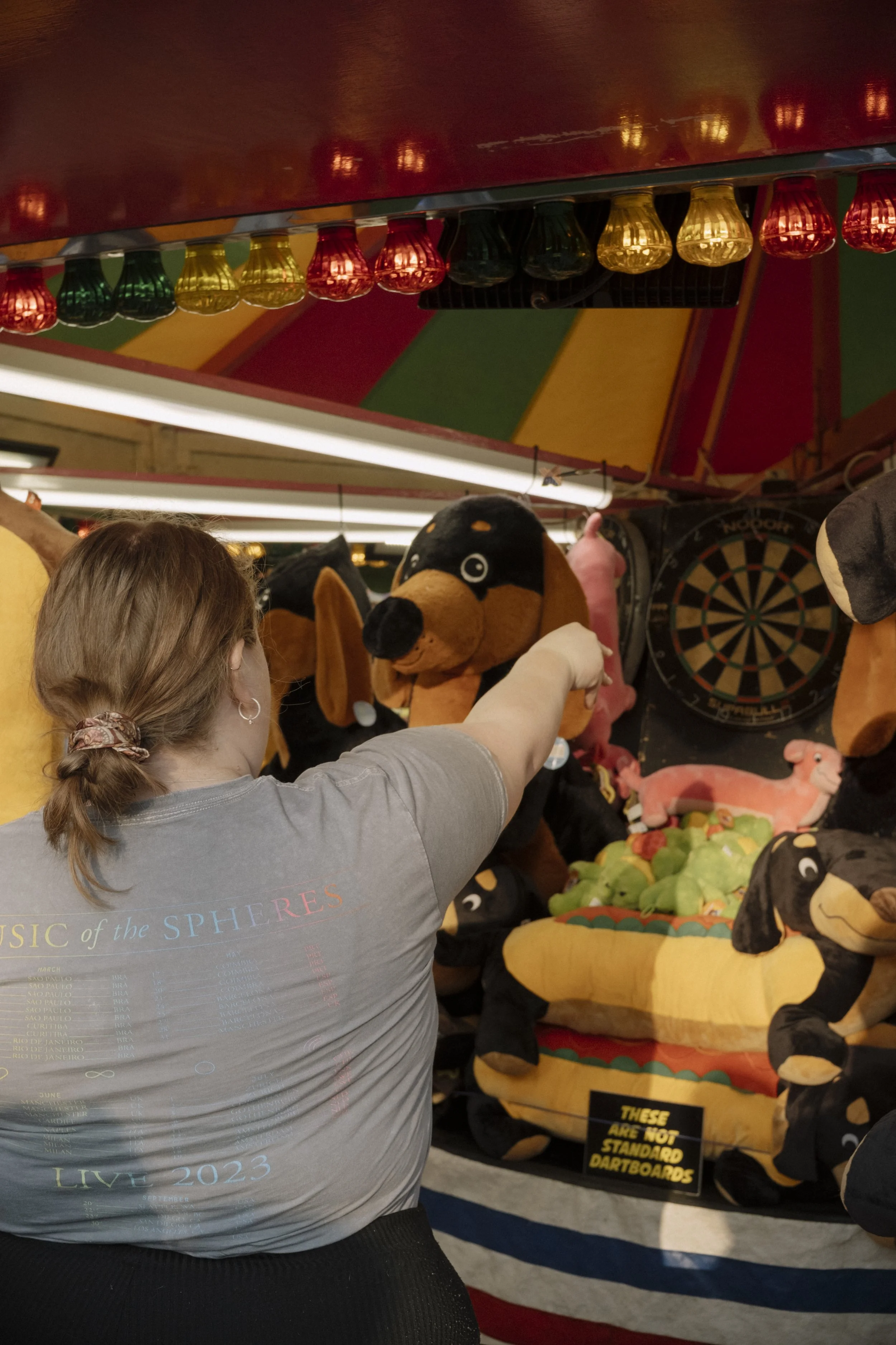 A woman with a braid wearing a gray t-shirt pointing at an arcade dartboard in a game area with plush animal prizes and colorful decorations.