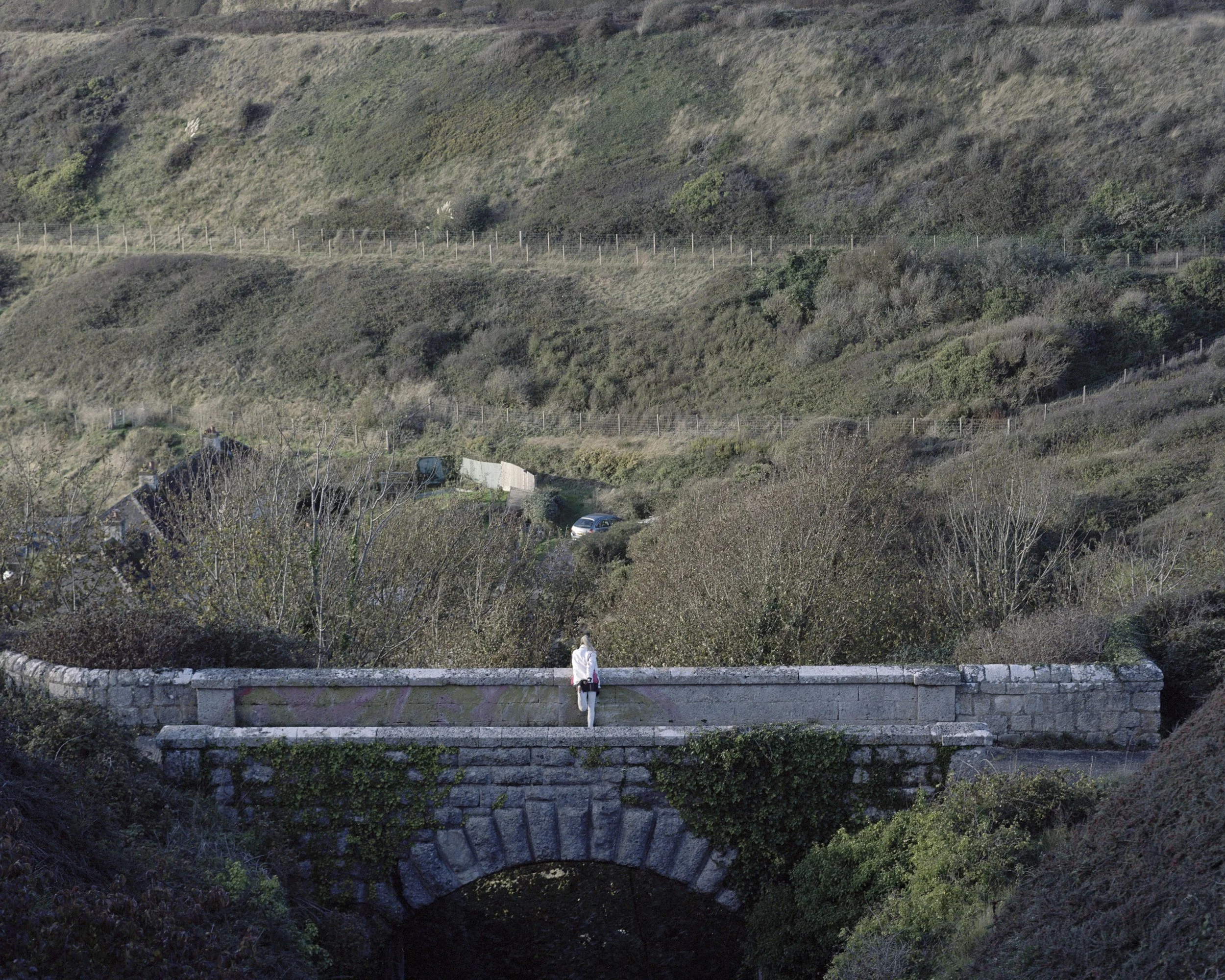 A person standing on a stone bridge in a hilly landscape with greenery, trees, and a few houses in the background.