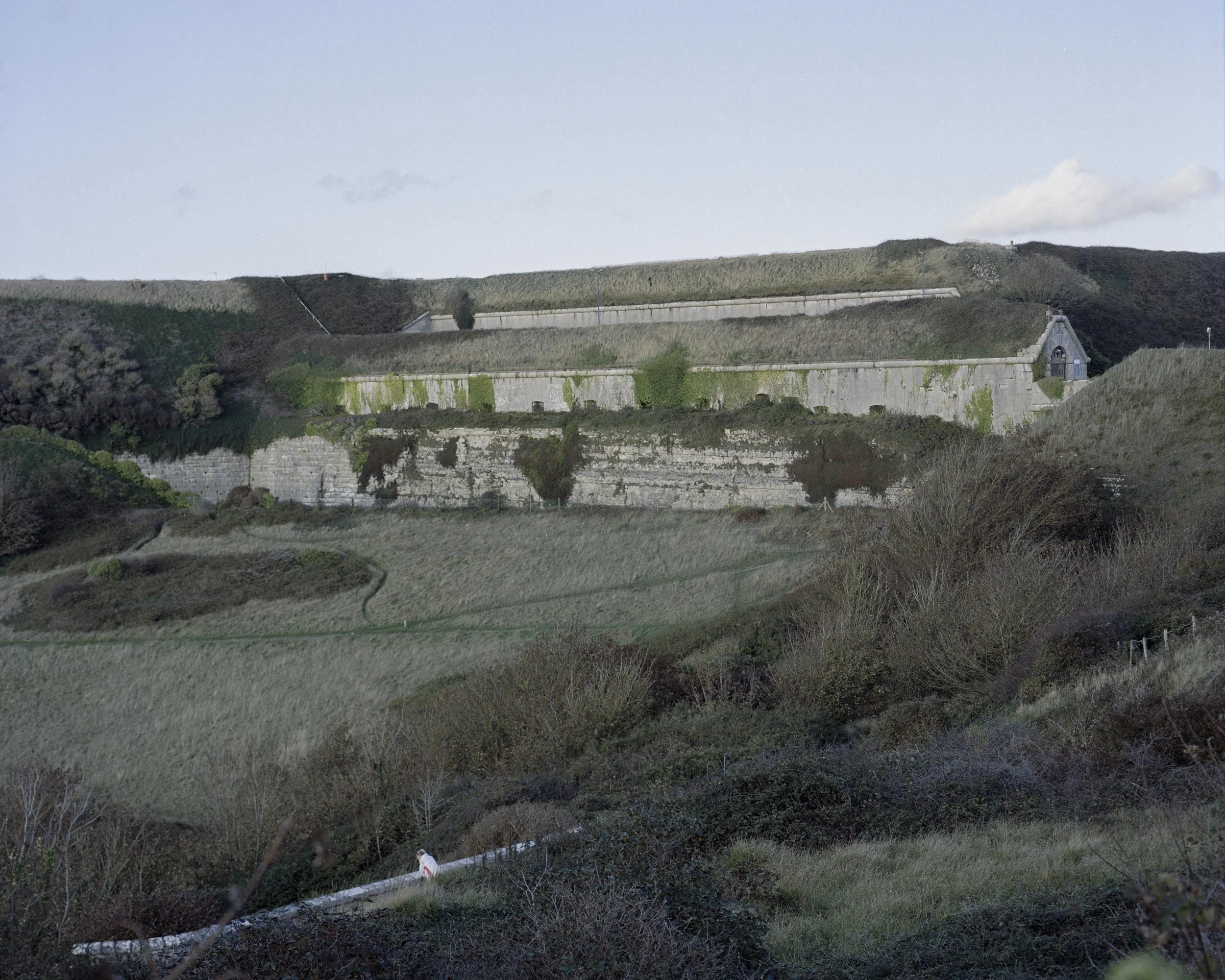 A landscape view of a hillside with green grass, shrubs, and a stone fortress with multiple levels and a small chapel or church at the top, under a partly cloudy sky.