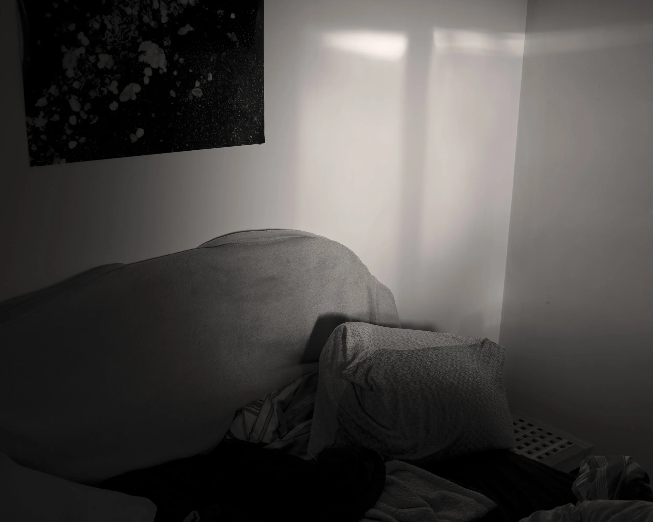 A dark bedroom with a bed covered in blankets and pillows, a white wall with a small painting, and a window casting light.