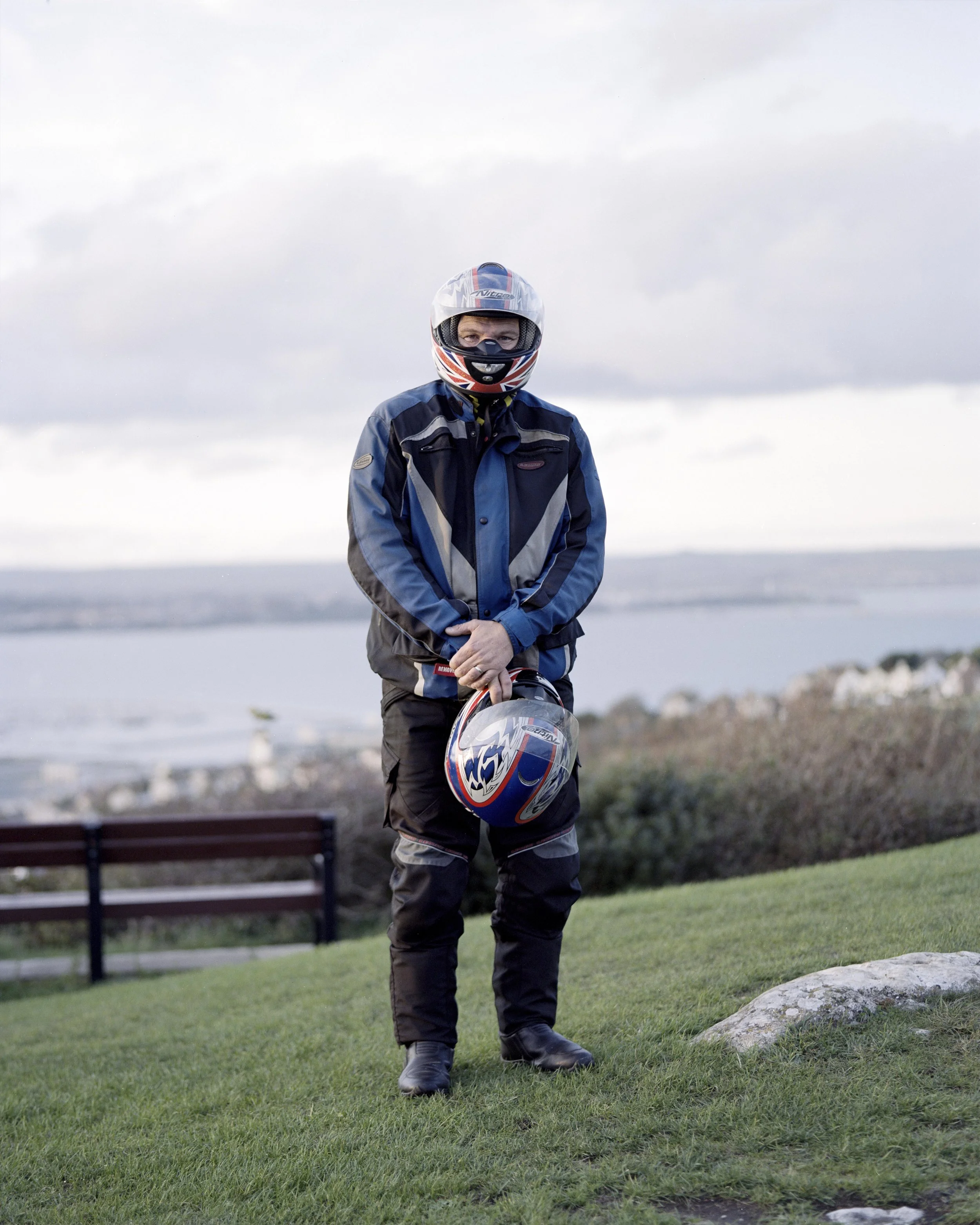 Person in motorcycle gear holding a helmet outdoors with water and landscape in the background.