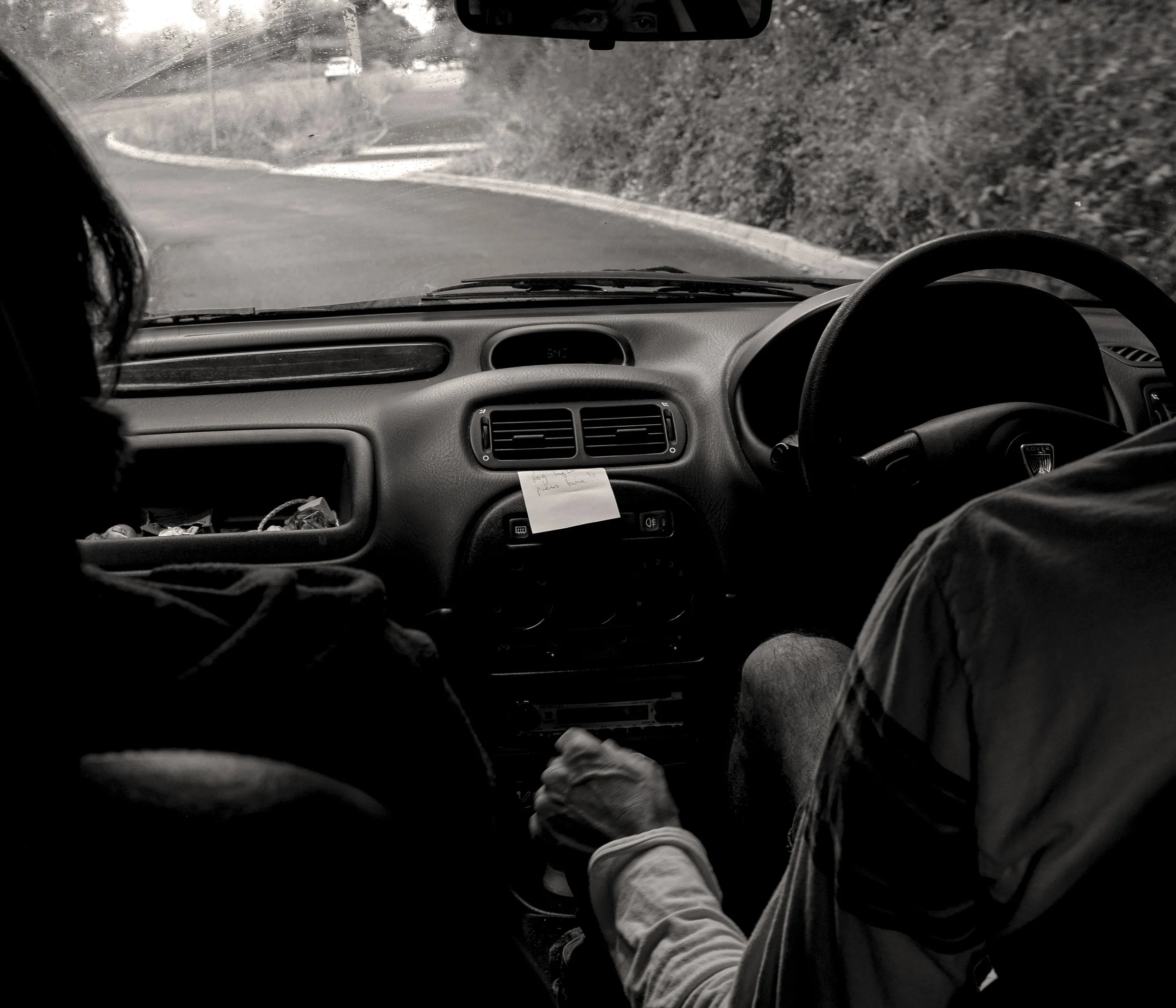 Inside a vehicle showing two passengers, the driver and one passenger on the left, in a rural area with trees blocking part of the view outside. The dashboard is visible with vents, knobs, and a note attached with some writing. The road ahead appears