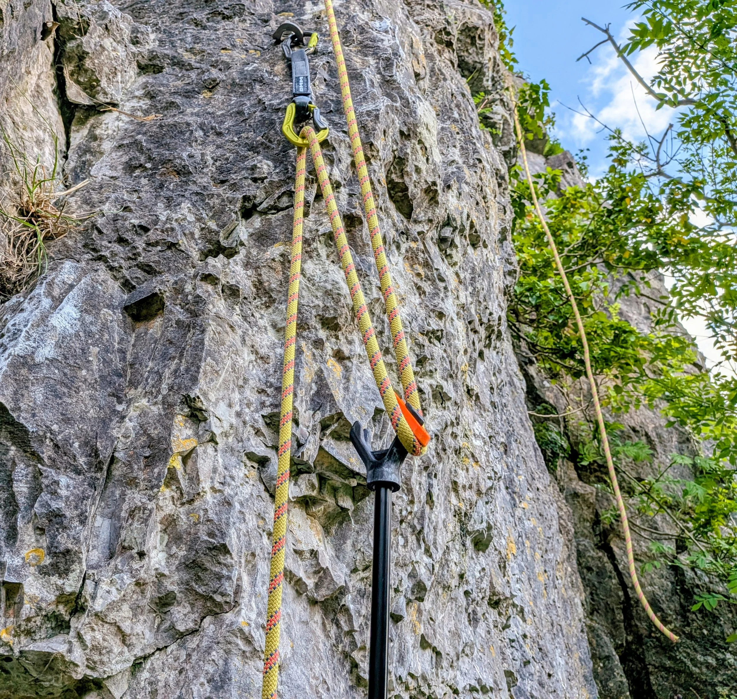 Close-up of climbing gear including a clipr clip stick and climbing ropes. The Black and orange clipr is hooking the rope.With a blue sky and green foliage in the background.