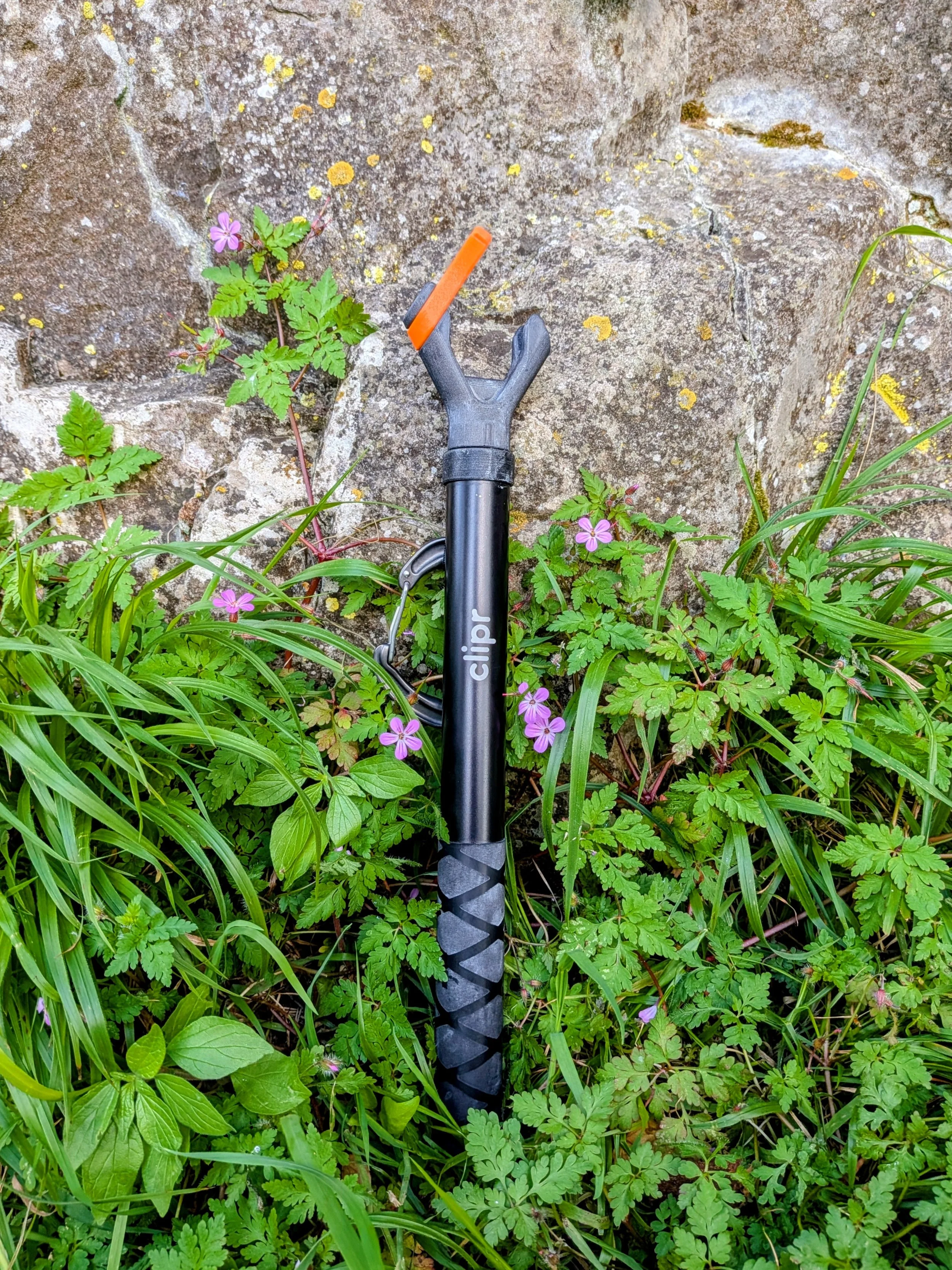 A black telescoping climbing clip stick pole with an orange clip resting on green grass and small purple flowers, against a mossy stone wall.