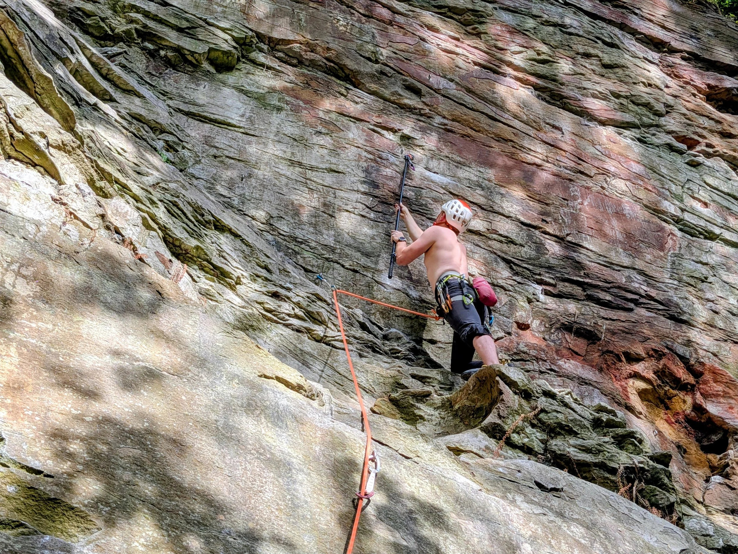 A shirtless man with a helmet and climbing gear is rock climbing on a steep, rugged cliff face. He is using a climbing harness and ropes for safety. He is using clipr to place a quickdraw.