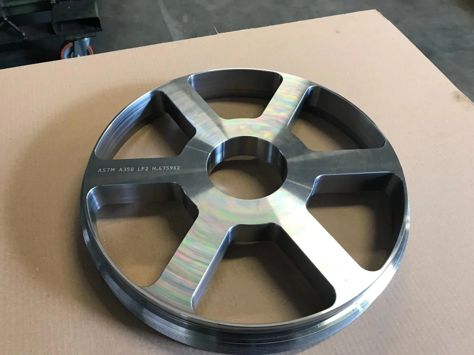 Ruota di acciaio lucido e spazzolato con marcature ASTM, posizionata su un cartone in un ambiente industriale.