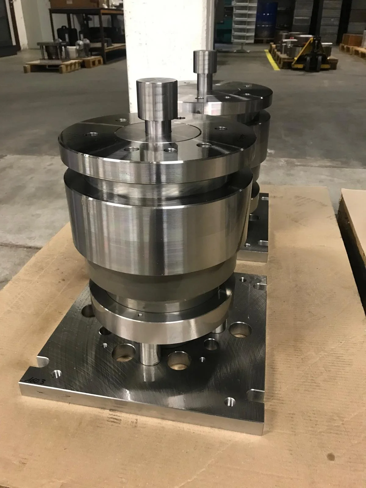 Un componente metallico industriale con parti cilindriche e piani piatti, posizionato su una base di metallo, probabilmente una macchina o uno stampo, in un ambiente di fabbrica o laboratorio.