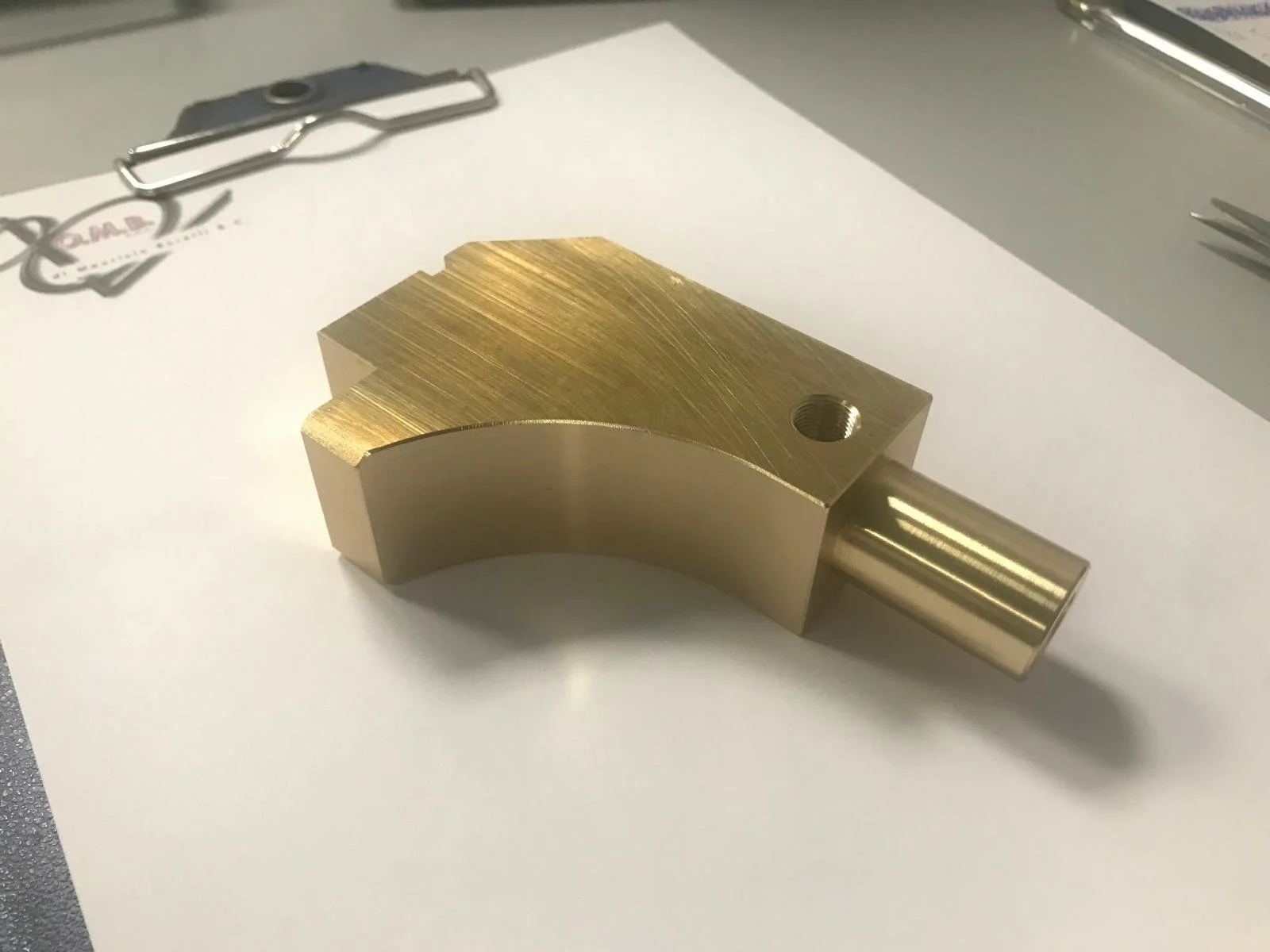 Componente meccanico in metallo dorato con una forma curva e un foro, posizionato su un foglio di carta bianca