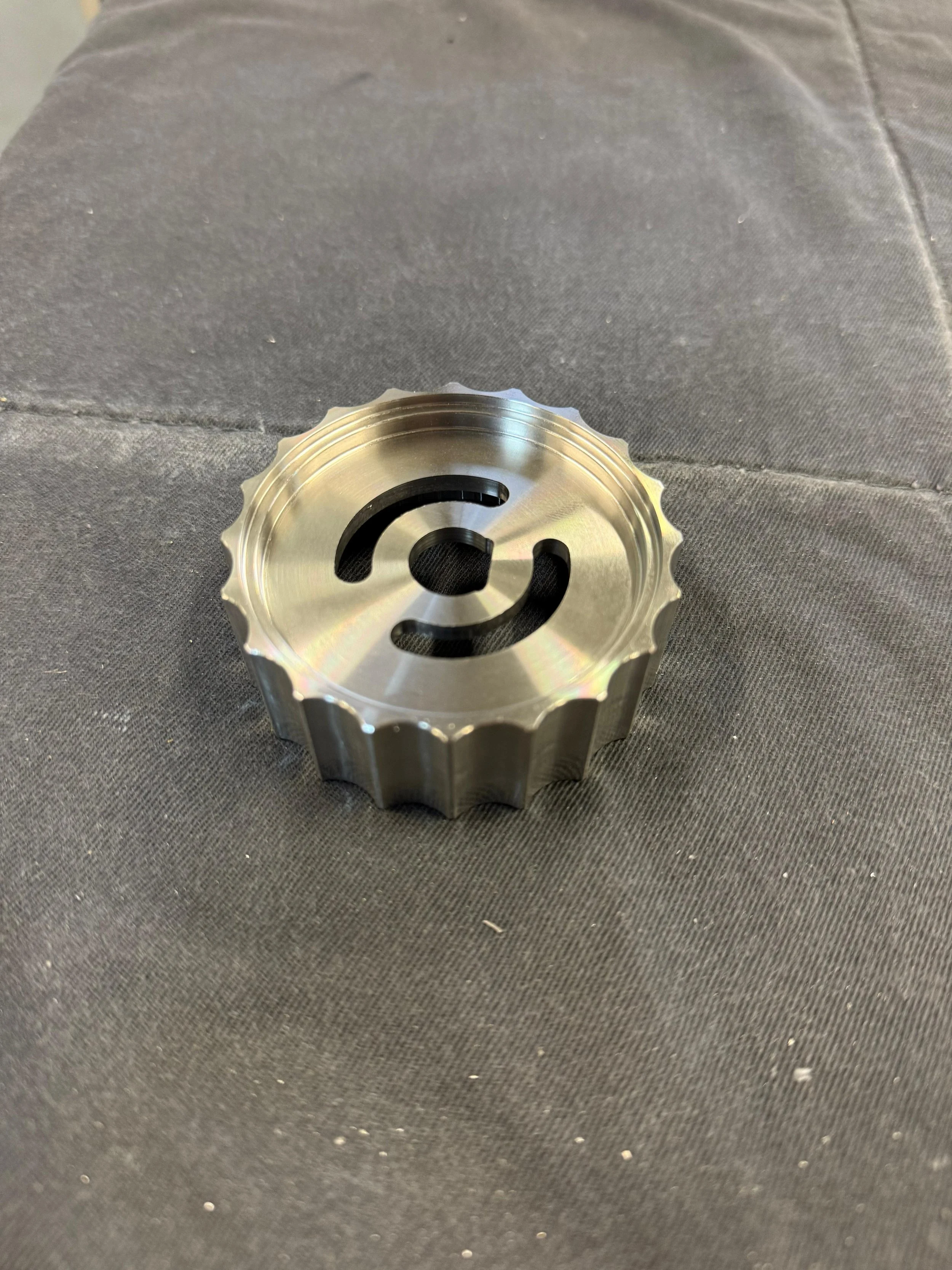 Pezzo meccanico in metallo con disegno a spirale al centro, posato su tessuto scuro.