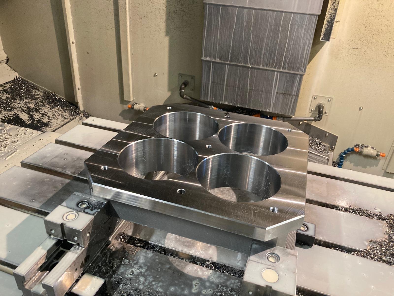 Un pezzo di metallo lavorato con fori rotondi, posto su una macchina utensile, circondato da trucioli metallici.