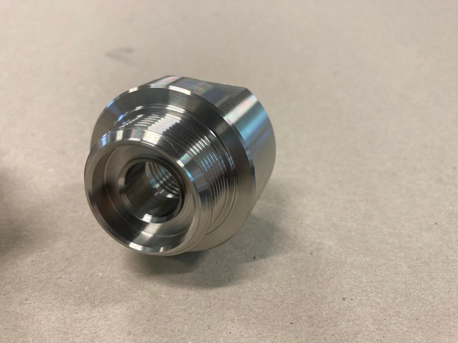 Component metallico cilindrico, lucido, con scanalature e foro centrale, su superficie liscia beige.