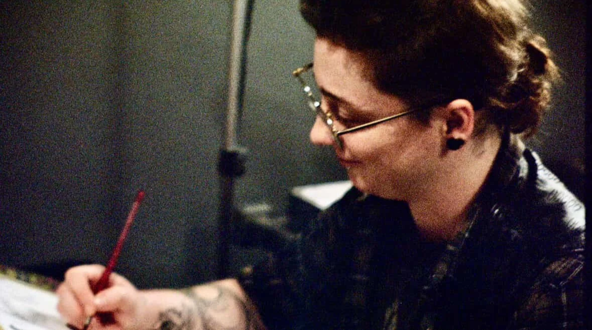 A person with glasses and tattoos on their arm, holding a red pencil, writing or drawing on a piece of paper in a dimly lit room.