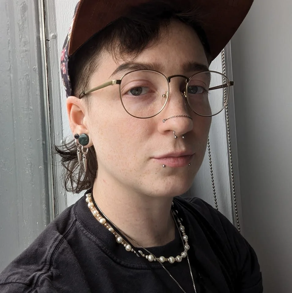 Close-up of a person with glasses, piercings, earrings, a pearl necklace, and a brown hat, standing near a window.