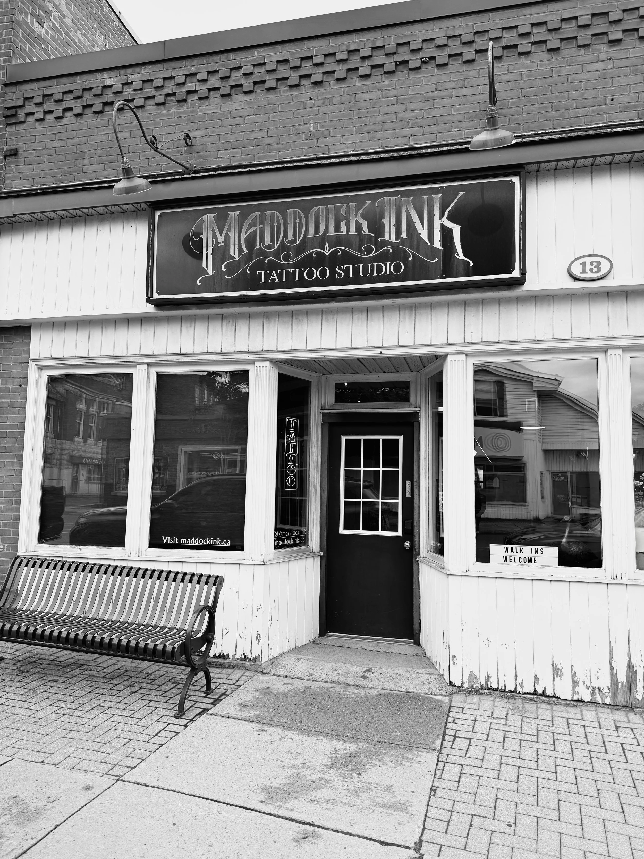 The front of a tattoo studio with a sign that reads 'Maddock Ink Tattoo Studio,' several large windows, a door, and a welcoming sign that says 'Walk Ins Welcome.' There is a bench outside on the sidewalk.