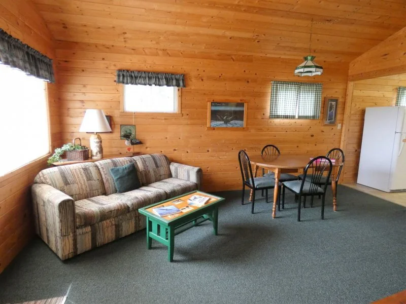 Living room with a striped sofa, green coffee table, round dining table with four black chairs, and a white refrigerator in a wood-paneled cabin.