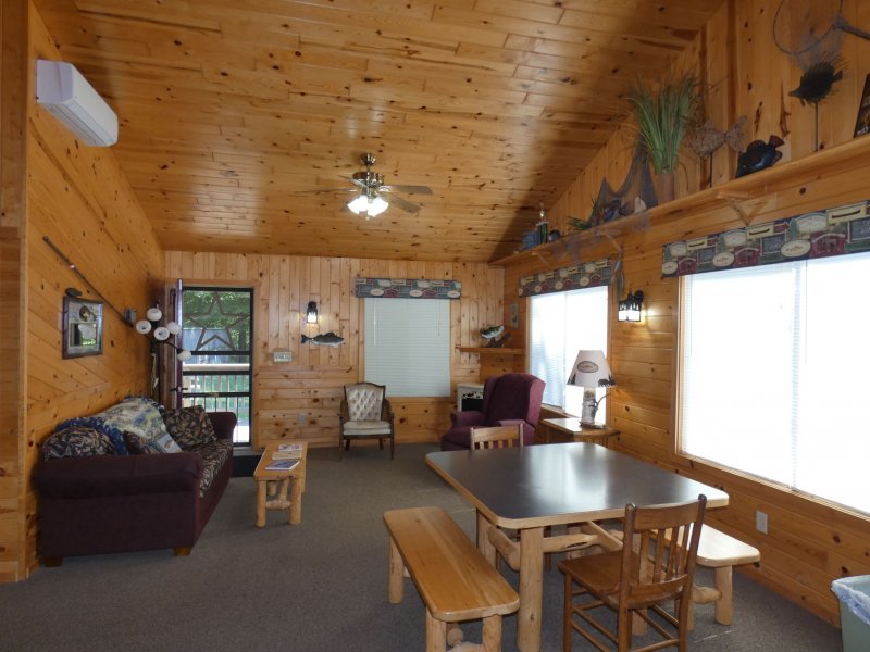 Wood-paneled living room with a sofa, two armchairs, a dining table with benches and chairs, windows with patterned valances, wall-mounted lights, an air conditioning unit, and a door leading outside.