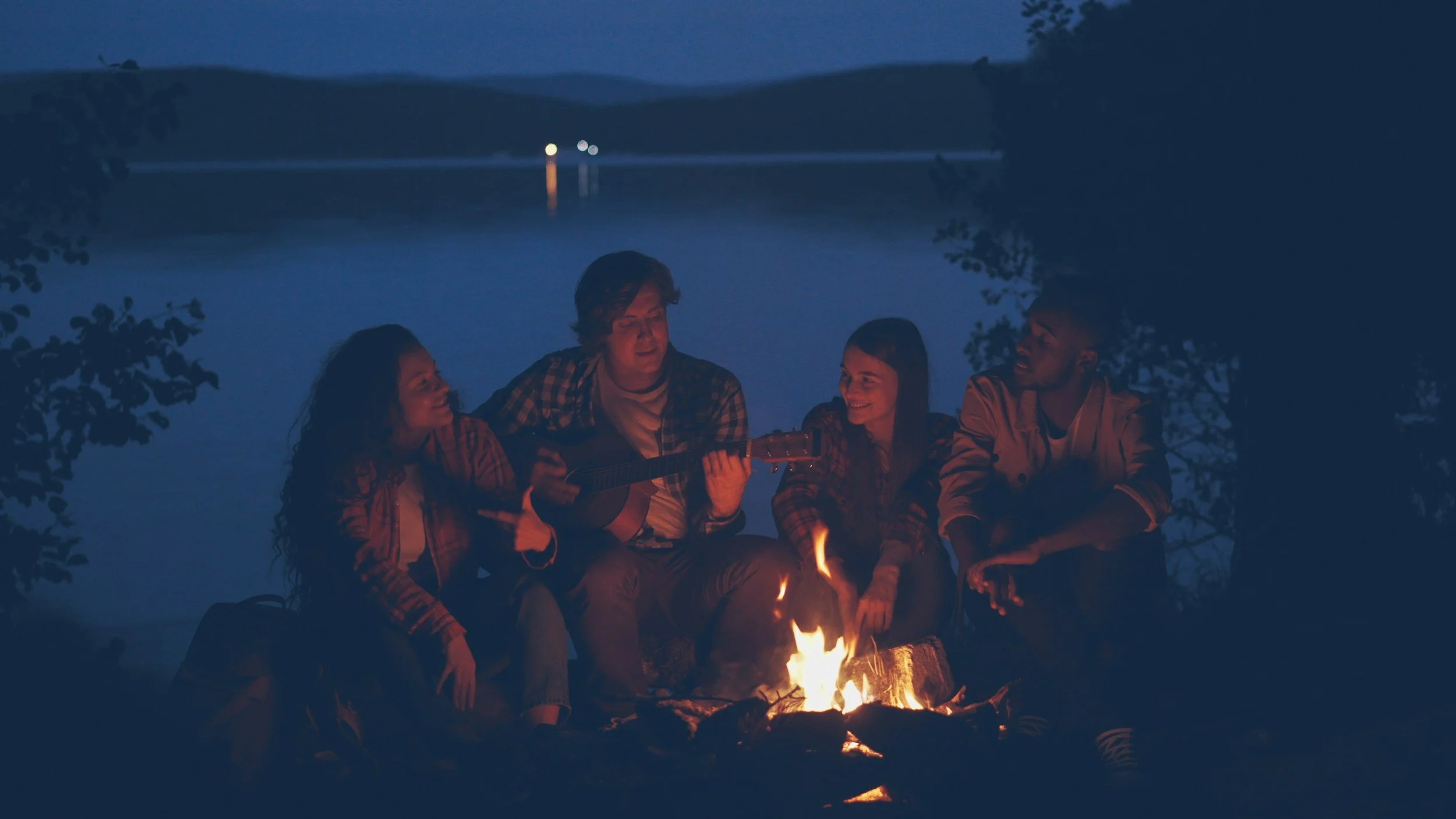 Group of five friends sitting around a campfire at night, with one playing guitar and others singing or chatting, with a lake and distant trees in the background.