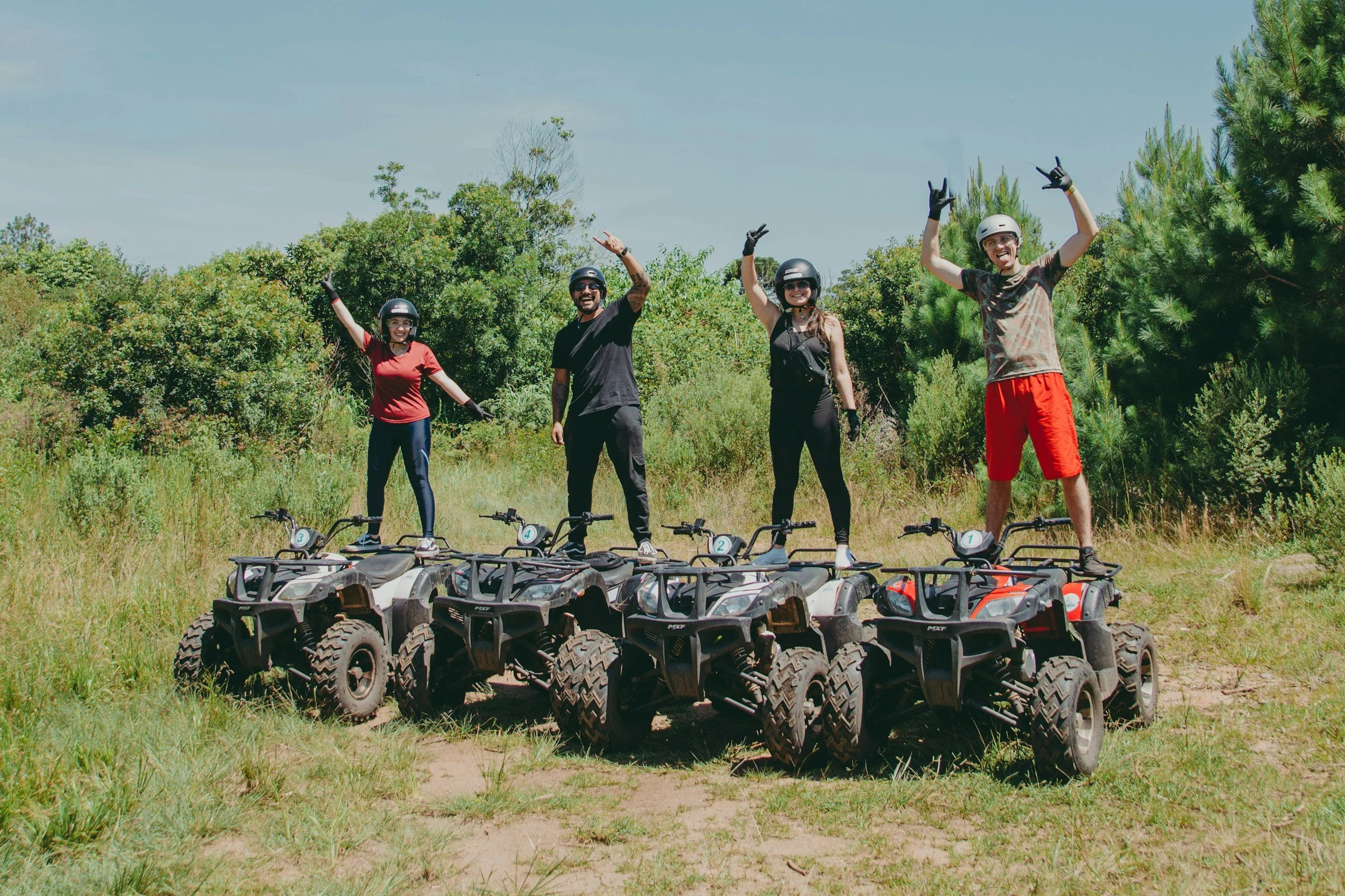 Four people wearing helmets and gloves standing on all-terrain vehicles (ATVs) with a row of ATVs on the ground in front of them, outdoors on grassy terrain with green bushes and trees in the background, under a clear blue sky.