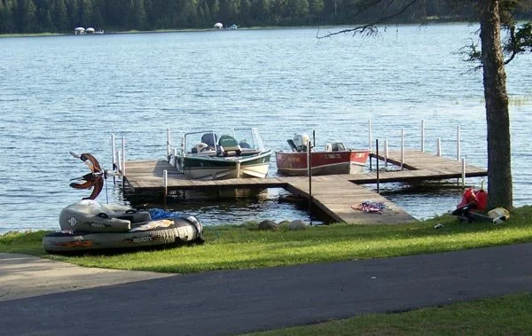 Dock with three boats and automatic inflator on the grass near a lake, with a small inflatable boat on the shore and a person sitting nearby.